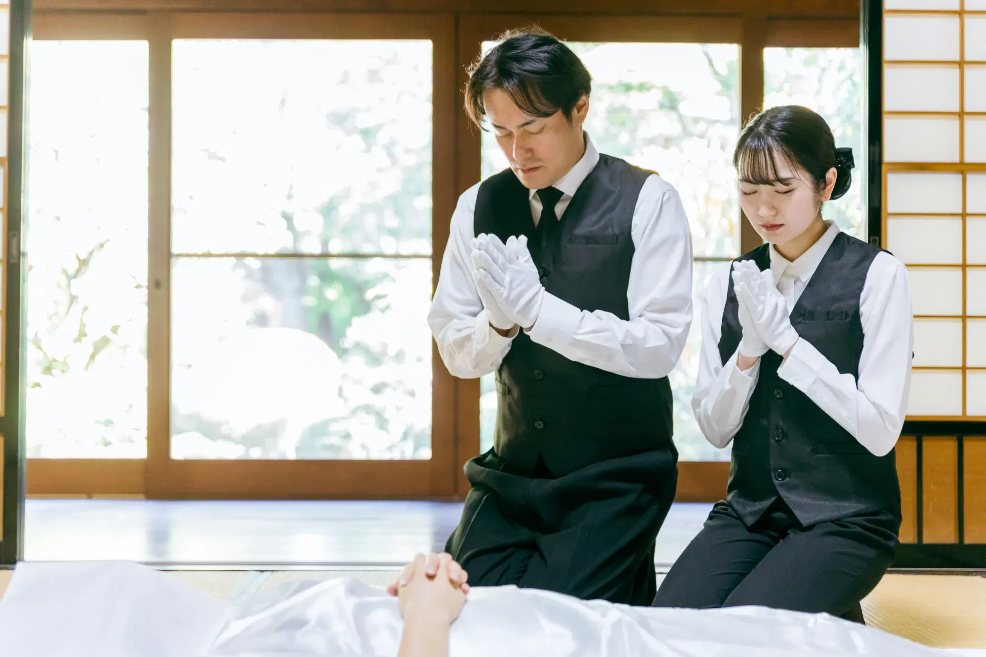 葬儀屋の男性と女性が遺体に手を合わせている。
