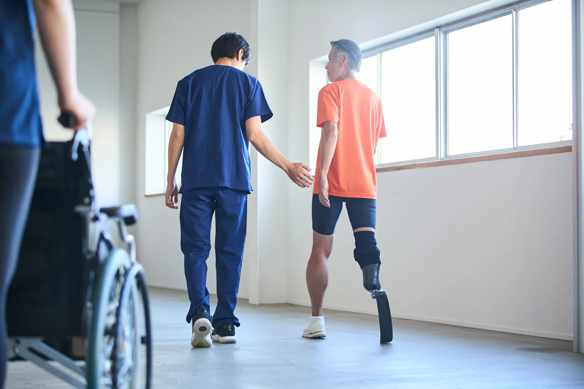 義足をつけて歩く練習をしているオレンジ色の服を着た男性と立って補助している青い服を着た男性