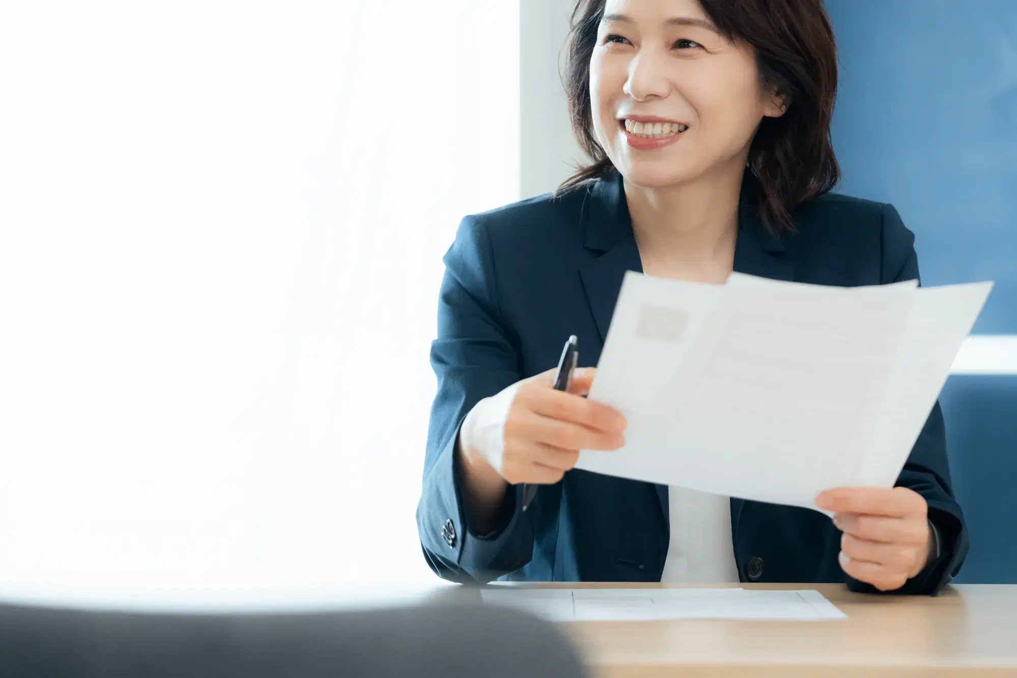 スーツの中年女性が右手にペンを持ち左手に2枚の資料を持って笑っている