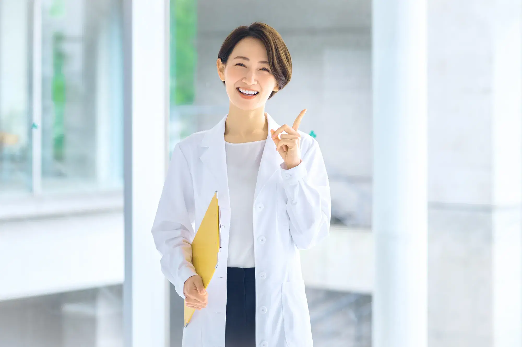 白衣を着て黄色いバインダーを持っている女性が笑顔で人差し指を立てている