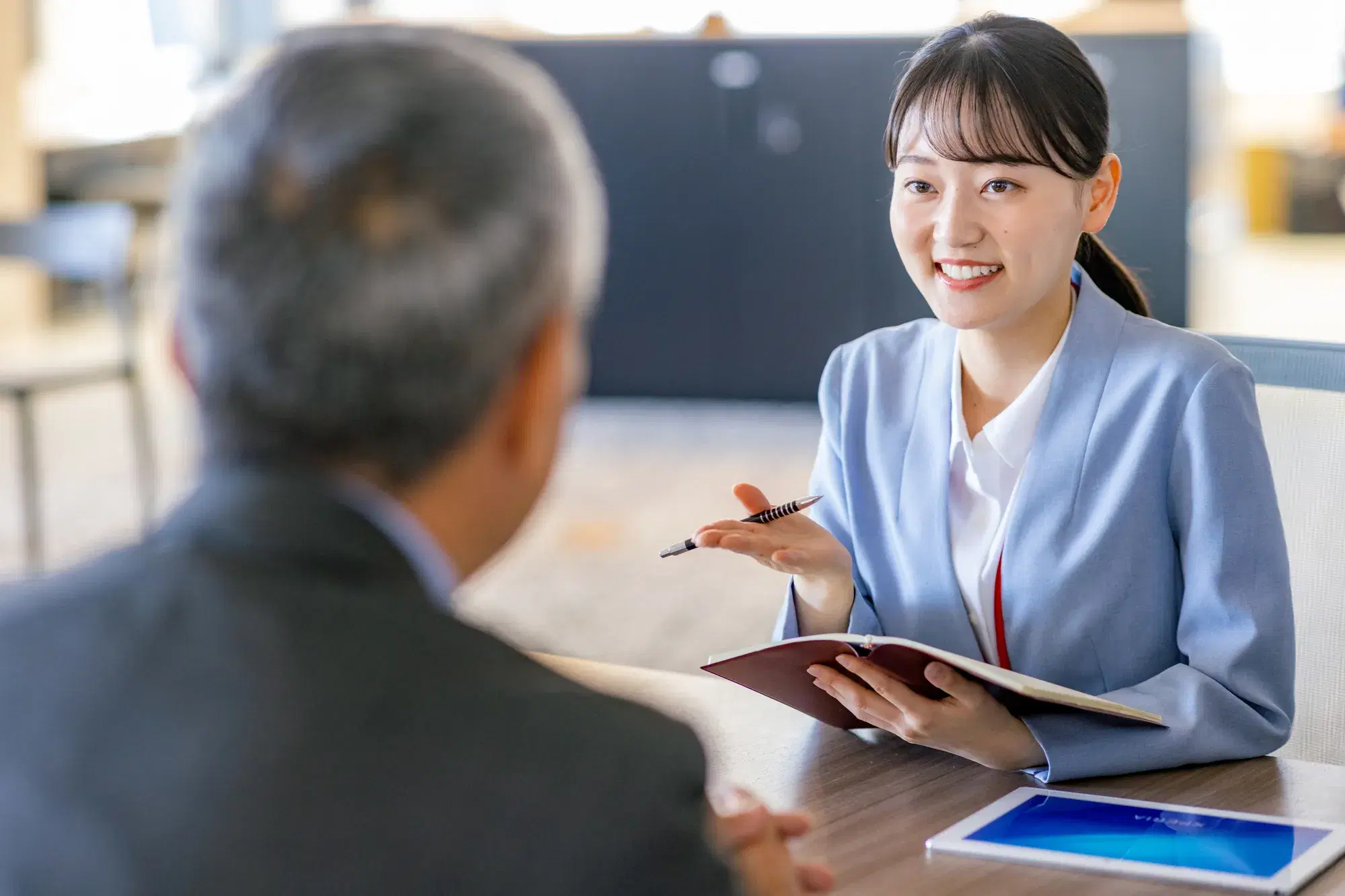 水色のスーツを着た女性がパンフレットを持って座りながら男性と会話