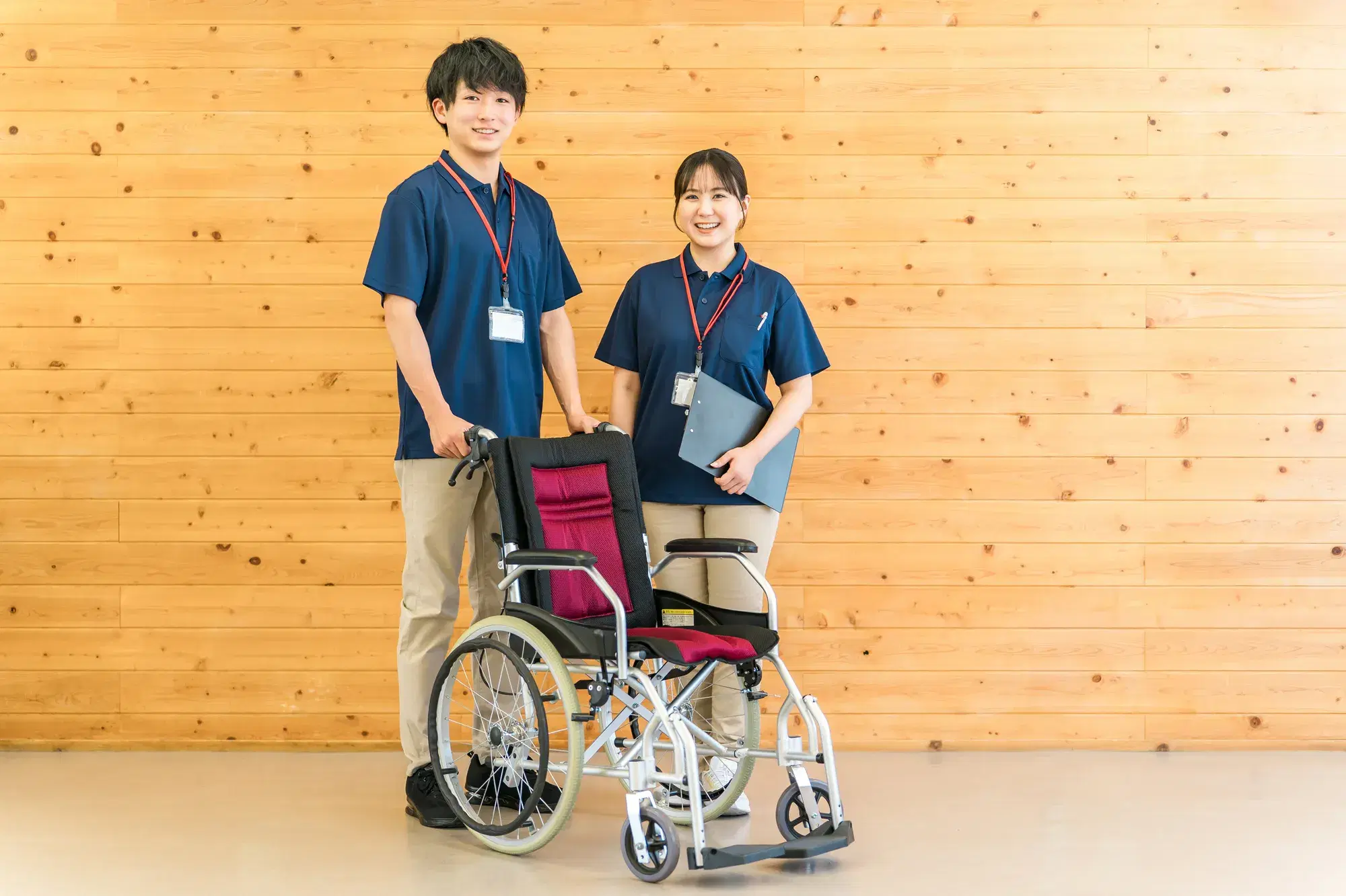 紺色の服を着ている医療従事者が二人おり、左の男性は車いすに手を置いており、右の女性はバインダーを持っている。