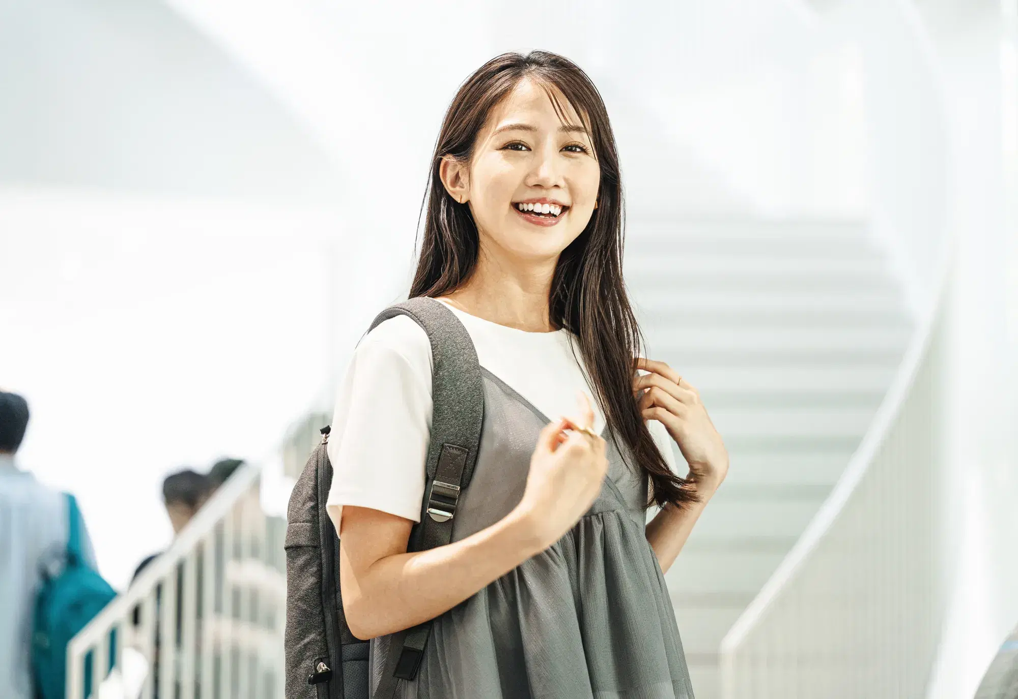階段の前で白と灰色のワンピースを着ておりリュックを背負っている女性が笑っている。