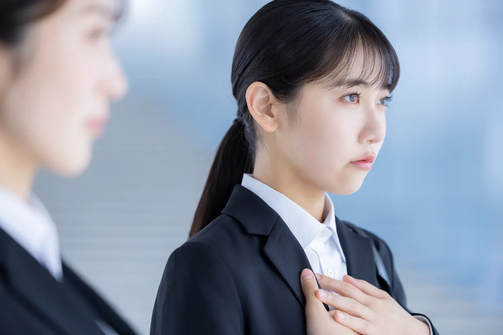 手前に黒いスーツを着た女性がおり、奥で黒いスーツを着た女性が胸の上に手を当てて横を向いている。