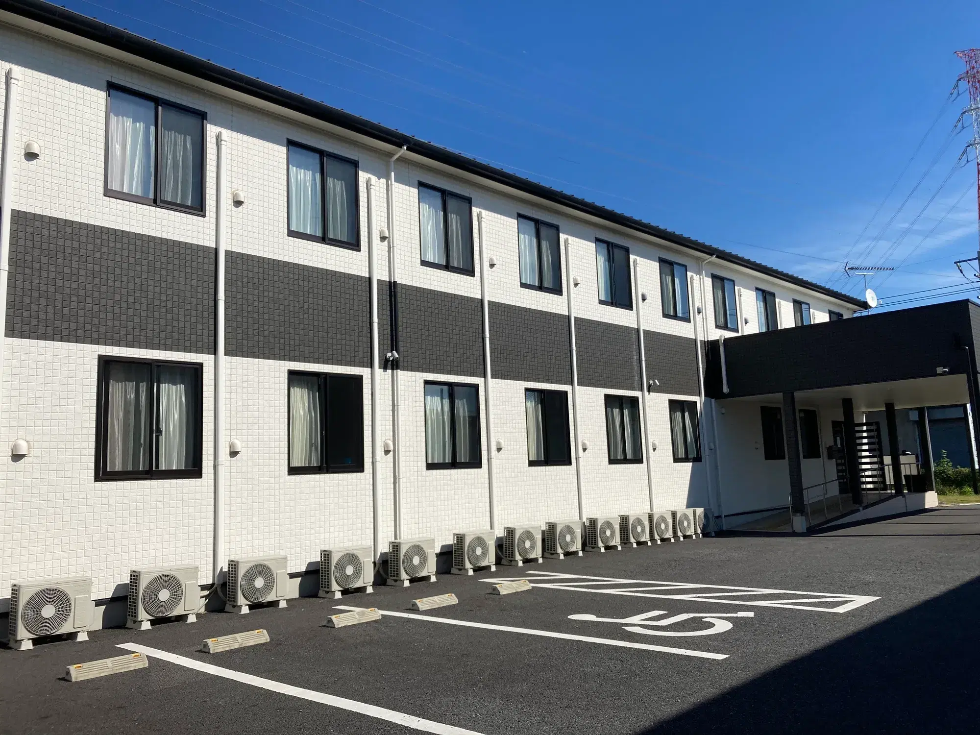 エアコンの室外機が並ぶ白と黒の建物と駐車場が写っている。