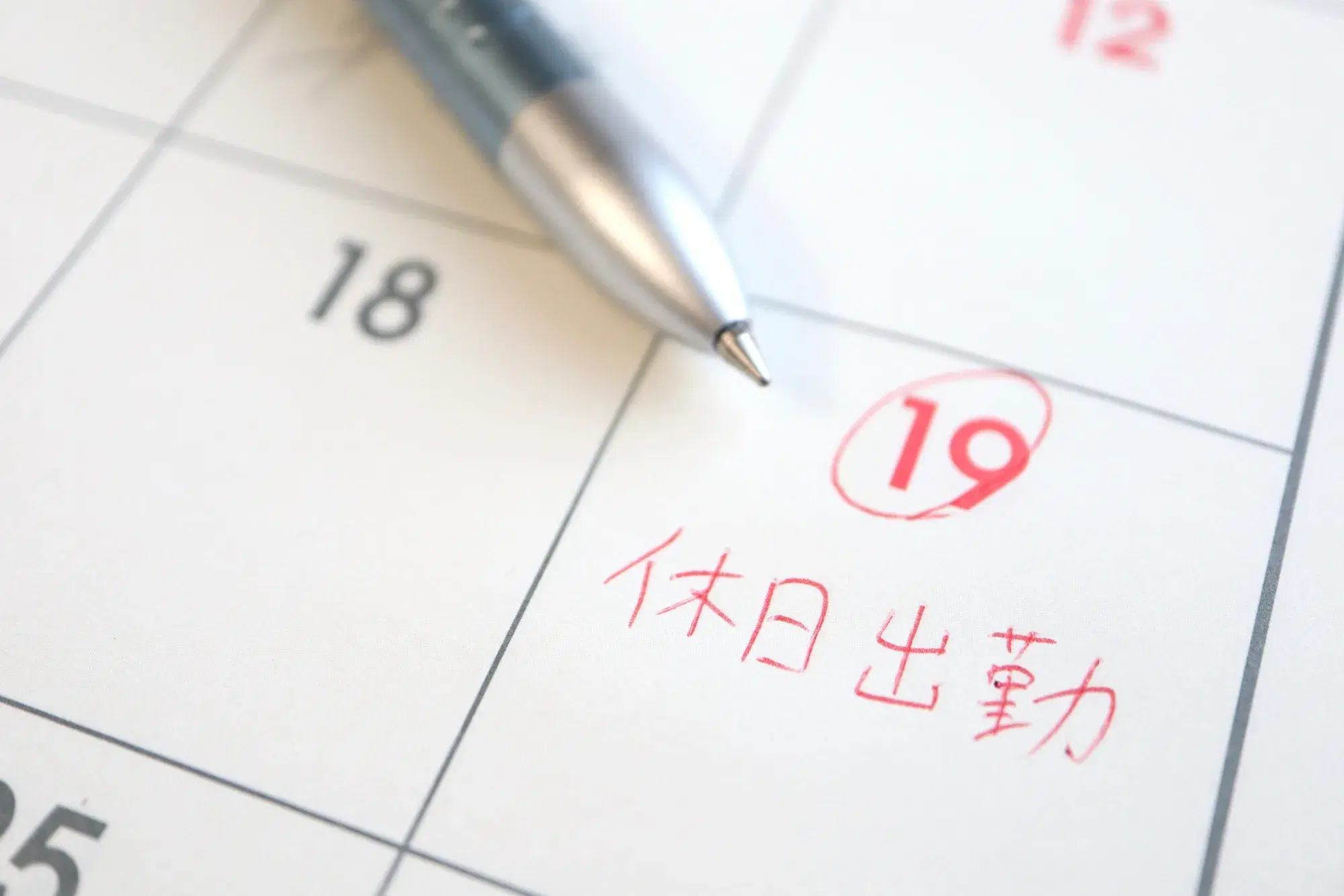 18日と19日が大きく映っているカレンダーの上にペンが置いてあり、19日には休日出勤と赤い文字で書いてある。