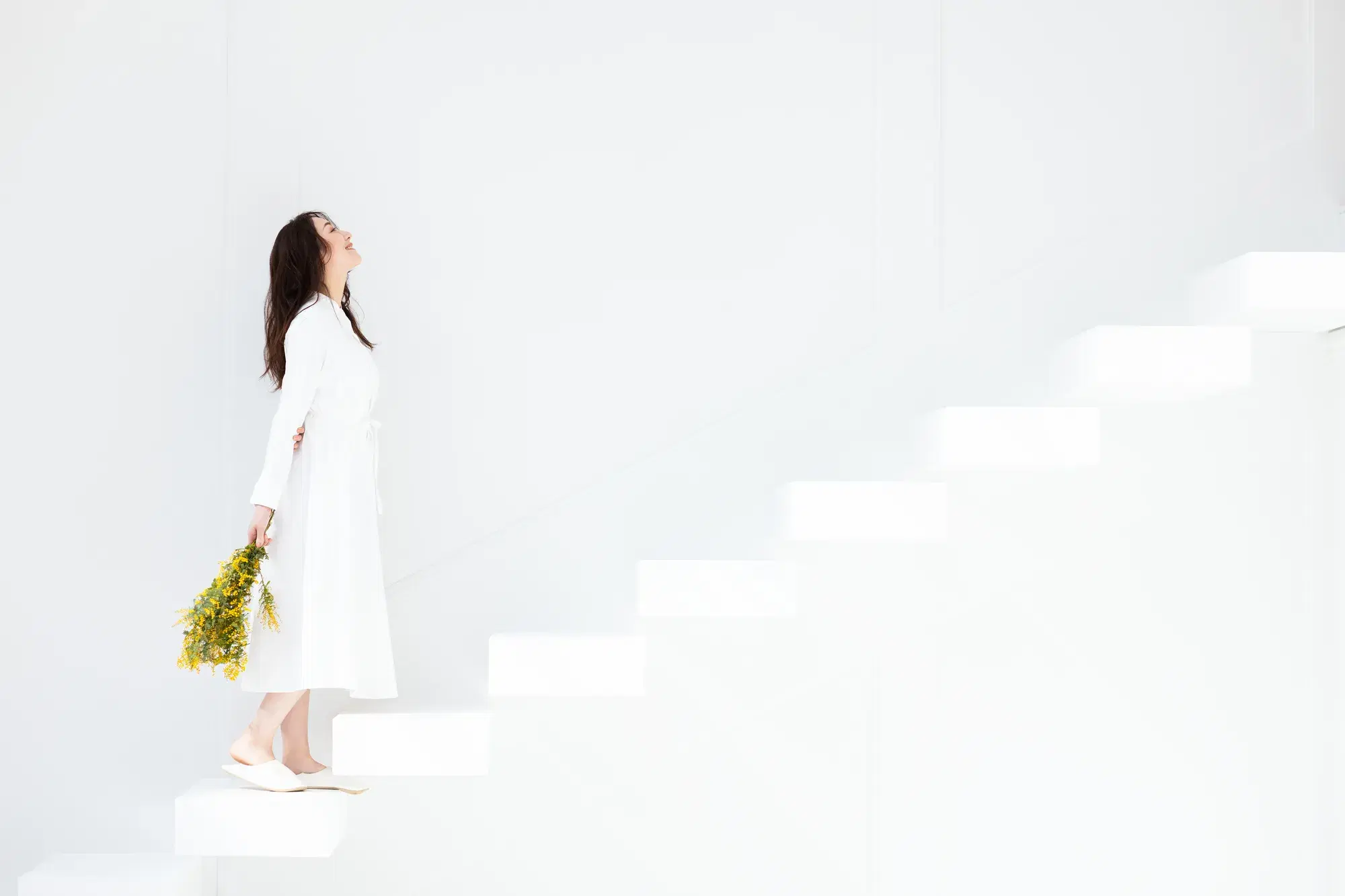 白いワンピースを着た女性が、黄色の花束を手に持って白い階段を上っている。