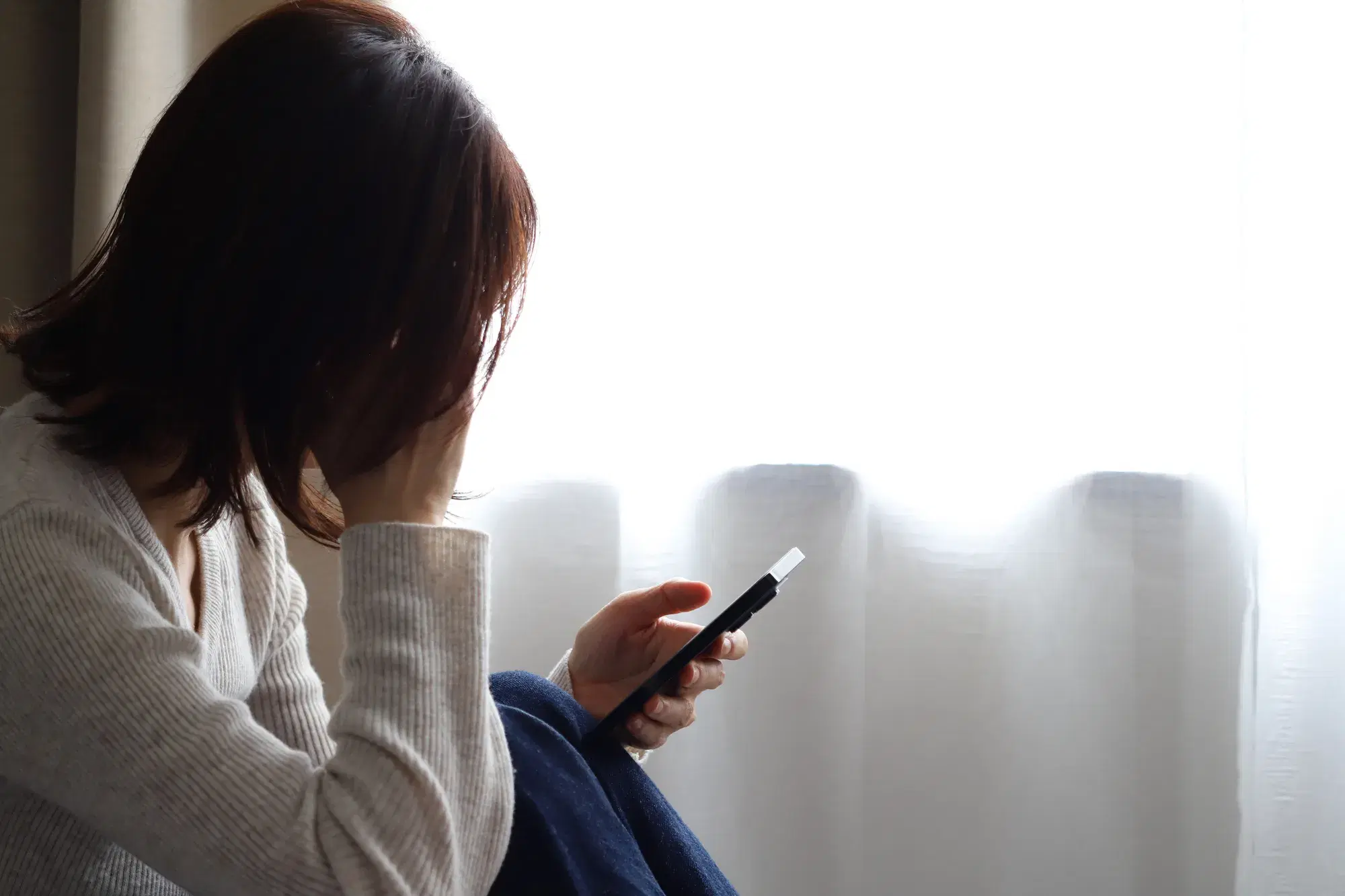 左手にスマホを持ち右手で頭を押さえている白い服を着た女性が座り込んでいる様子