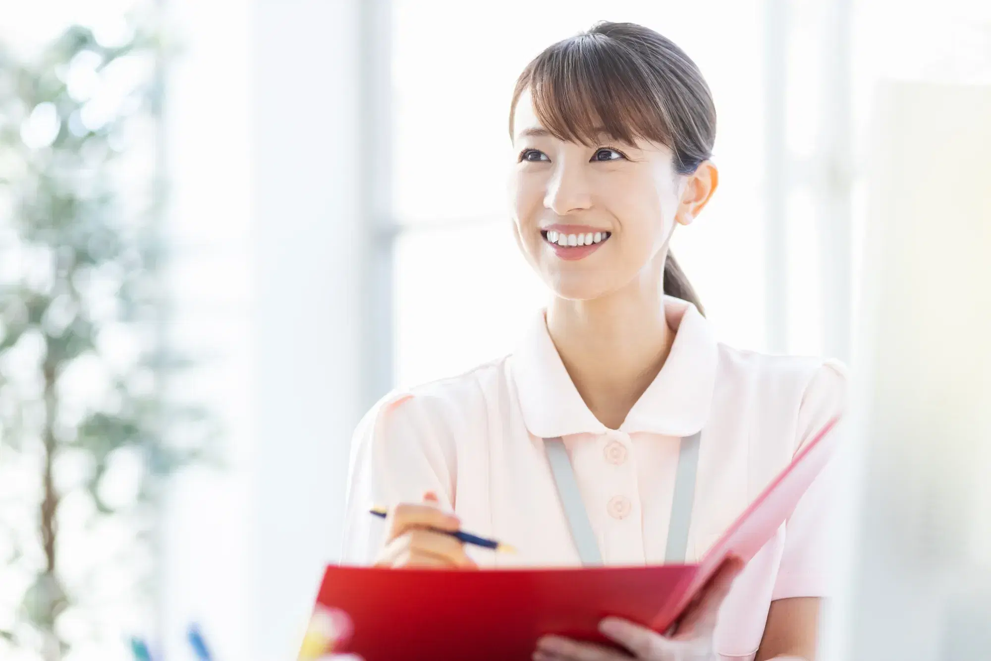 ピンク色の服を着た女性が赤いバインダーをに筆記用具で記入
