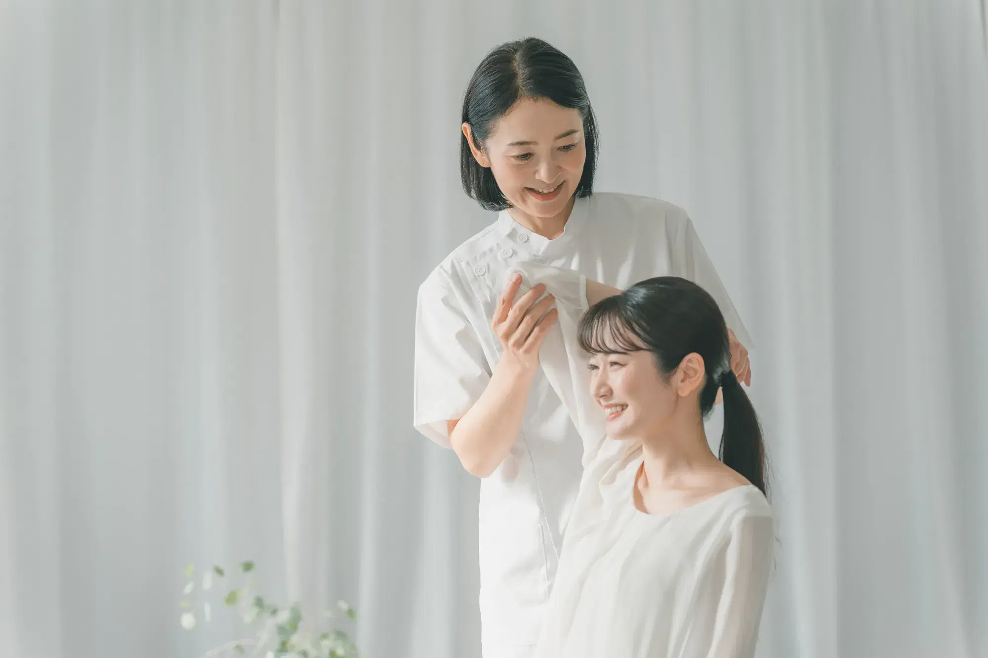白い服を着た女性2人のうち片方が椅子に座り肩肘を耳の位置まであげ、片方がその肘を支えている
