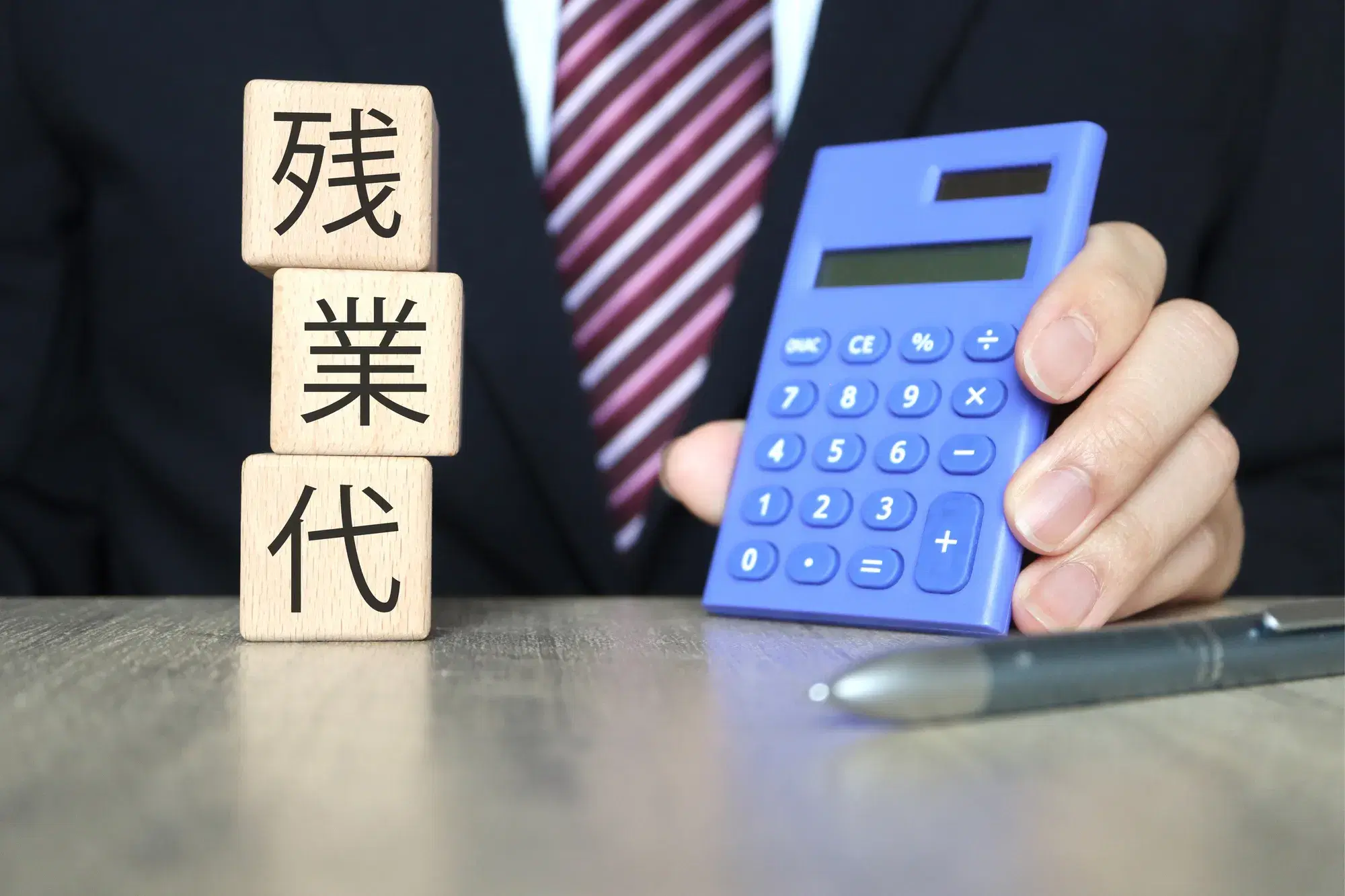 黒いスーツを着た男性が青い電卓を持っており、手前に「残業代」と書かれた木製ブロックとペンが置かれている様子が写っている。