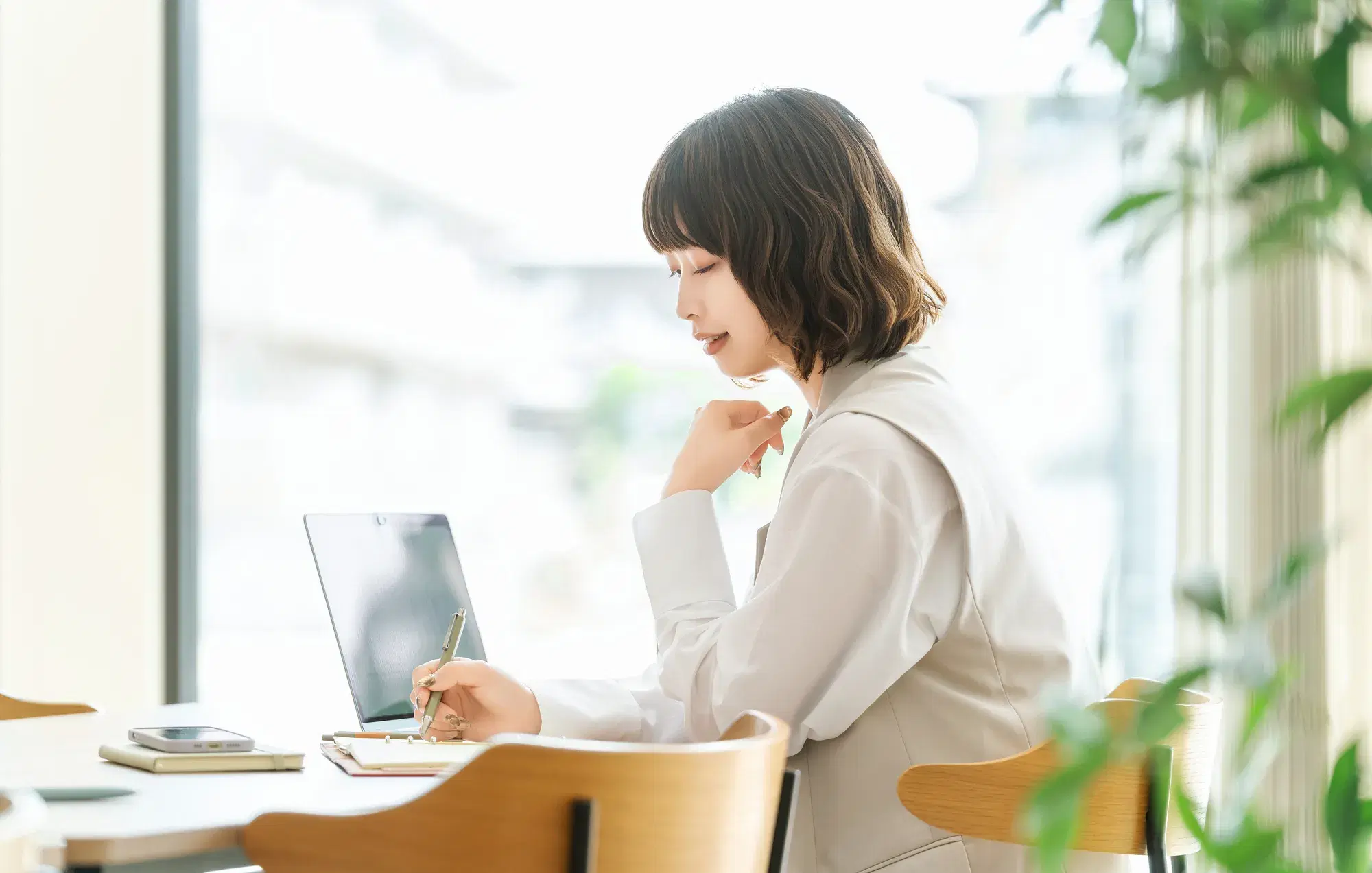 短い髪をしており白い服を着た女性がパソコンとノートと筆記用具を用いて勉強をしている