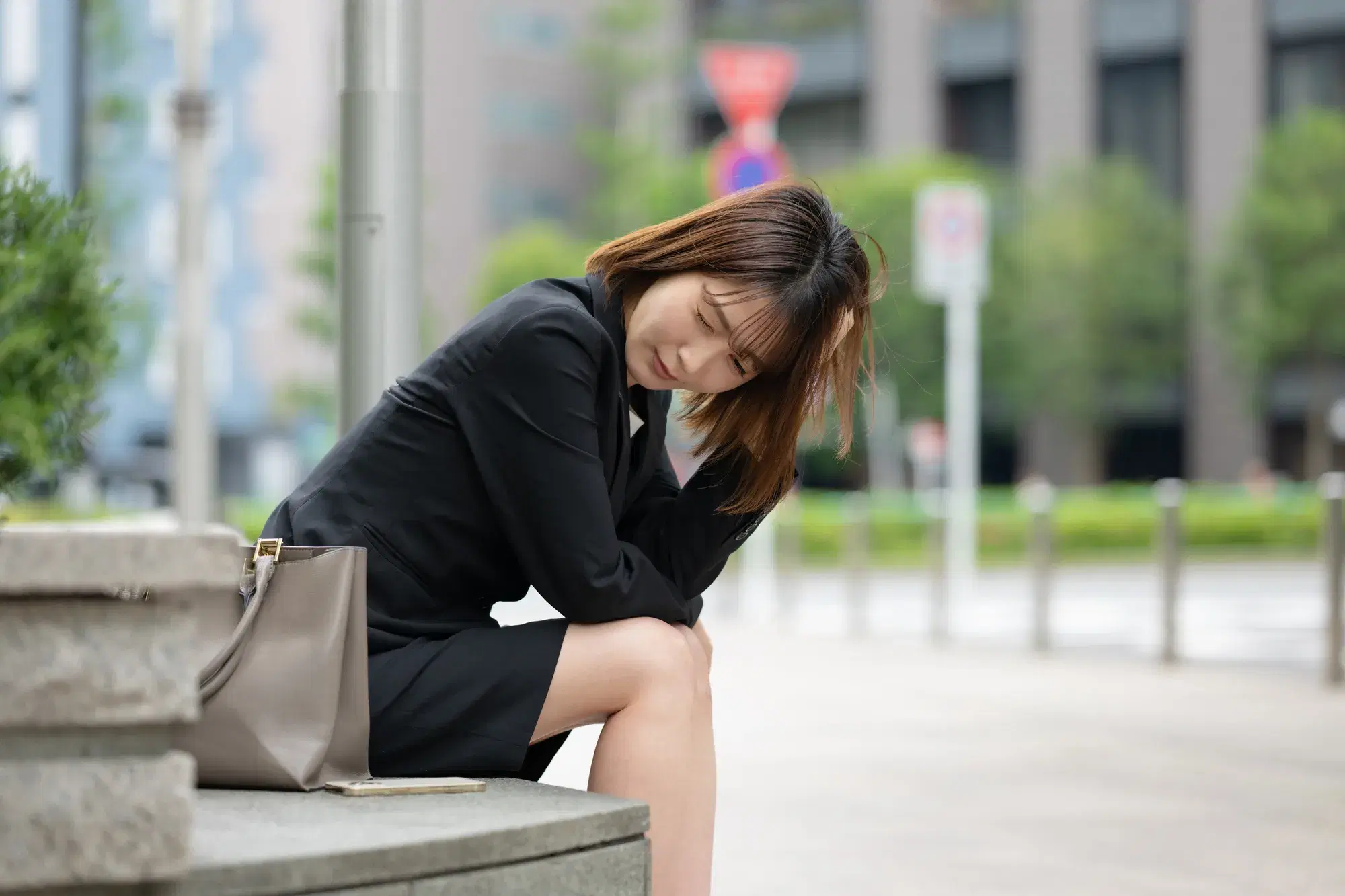 黒色のスーツを着た女性がベンチに座って頭を抱えている