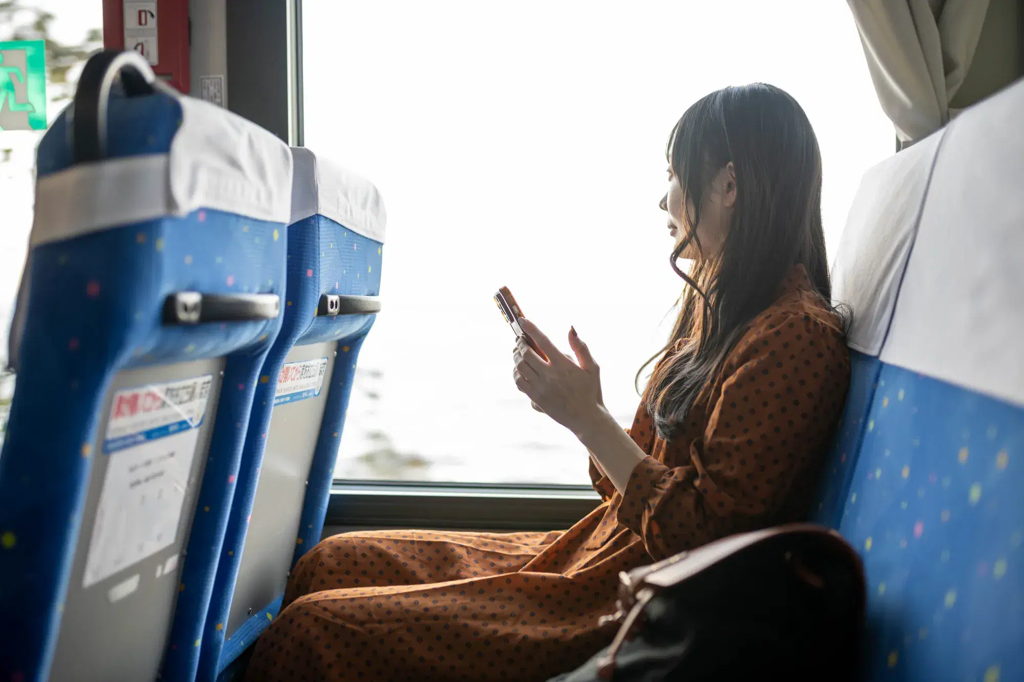 茶色のワンピースを着た女性がバスに乗りながらスマホを操作している。