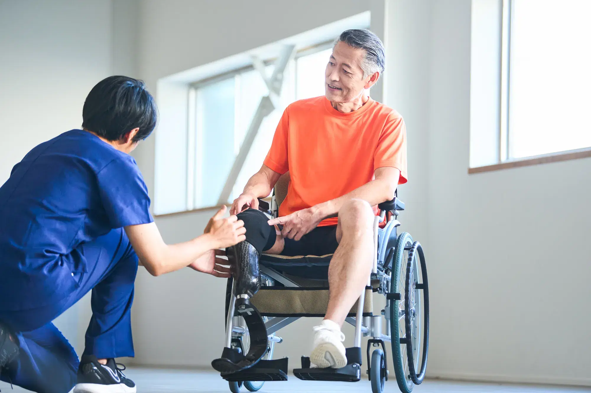 車いすにオレンジ色の服を着た義足を付けた男性と援助する青い服を着た男性