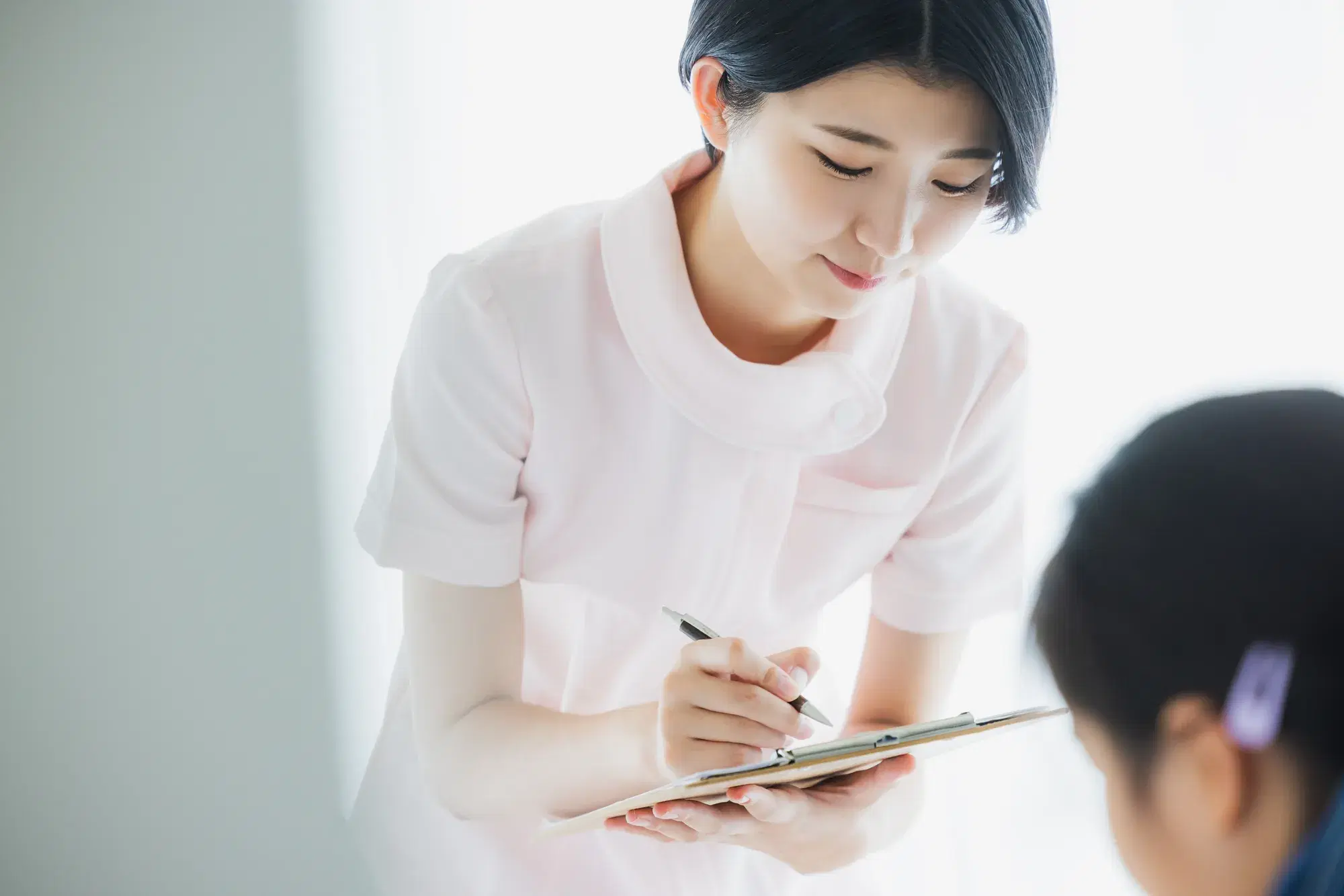 ピンクの医療用の服を着た女性が目の前にいる患者の子供の話を聞きながらバインダーに挟んだ書類に書き記している