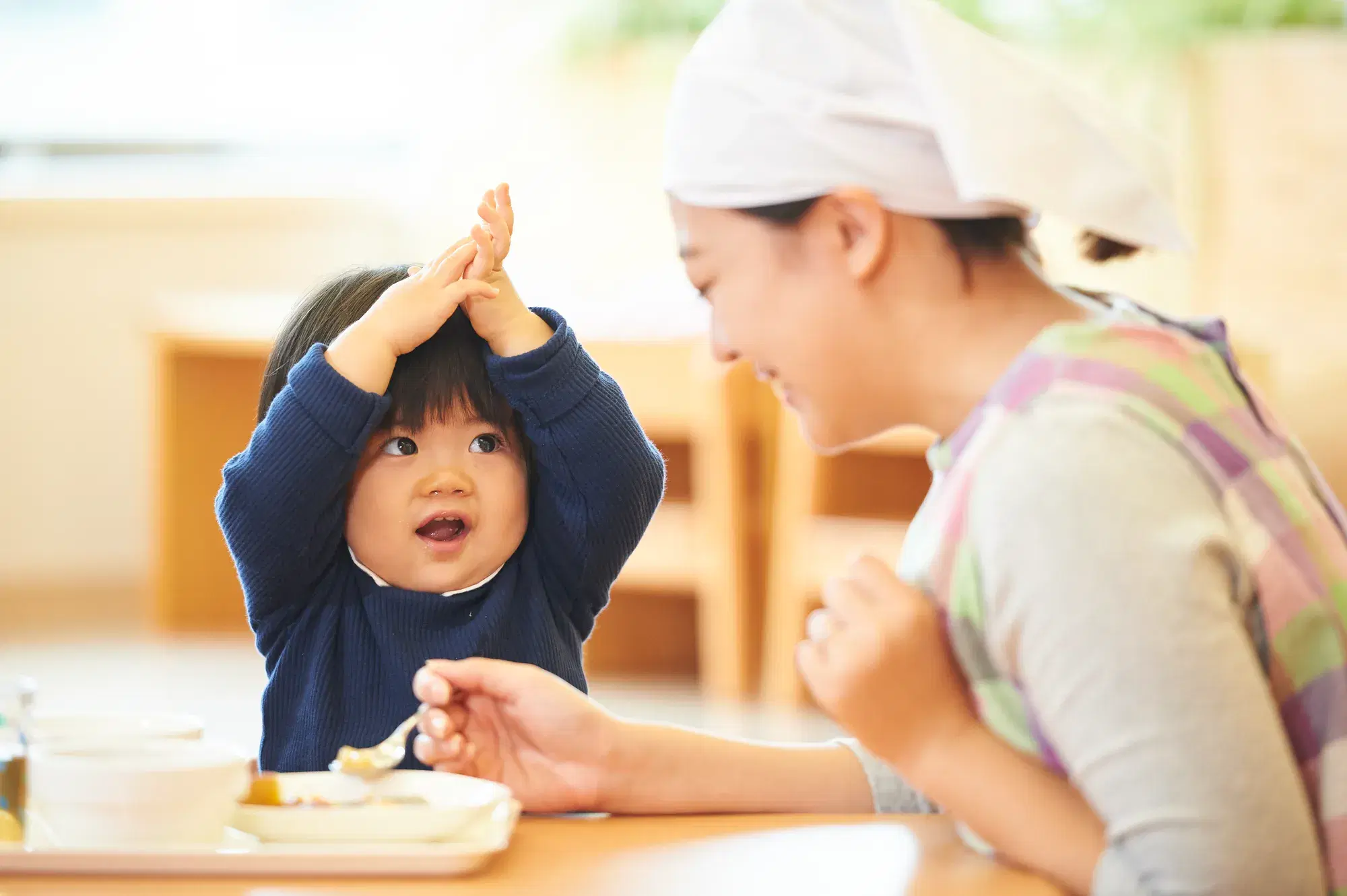 青い服を着た子どもが手を合わせながら腕を上げていて、それを笑顔で対応しながらご飯をあげる女性