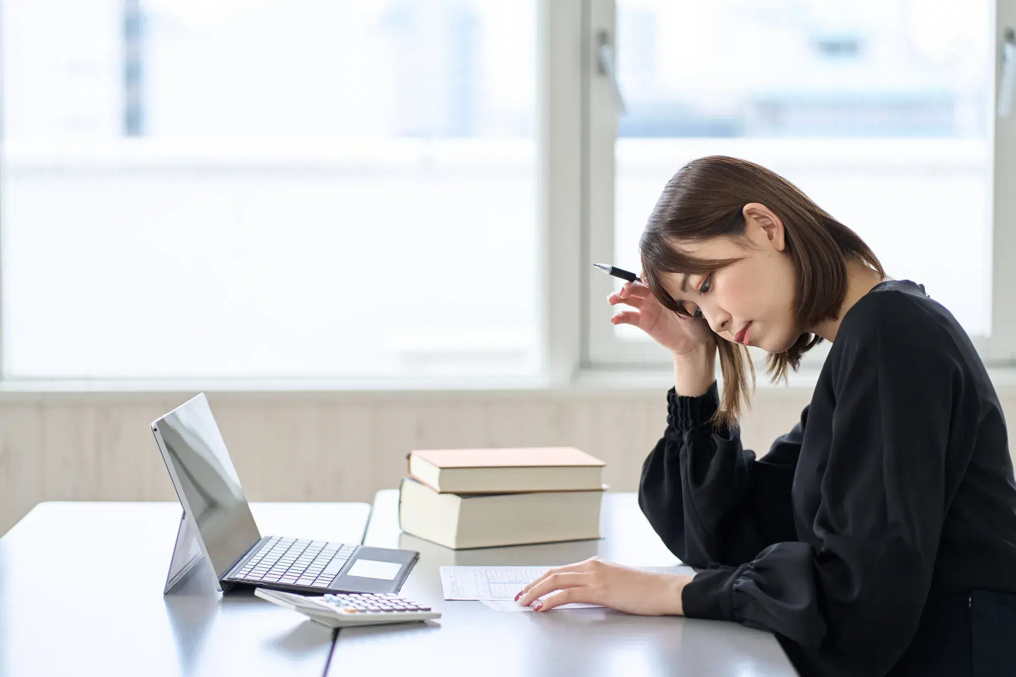 黒い服を着た女性が筆記用具を持った右手を頭に添えながら悩んでいる