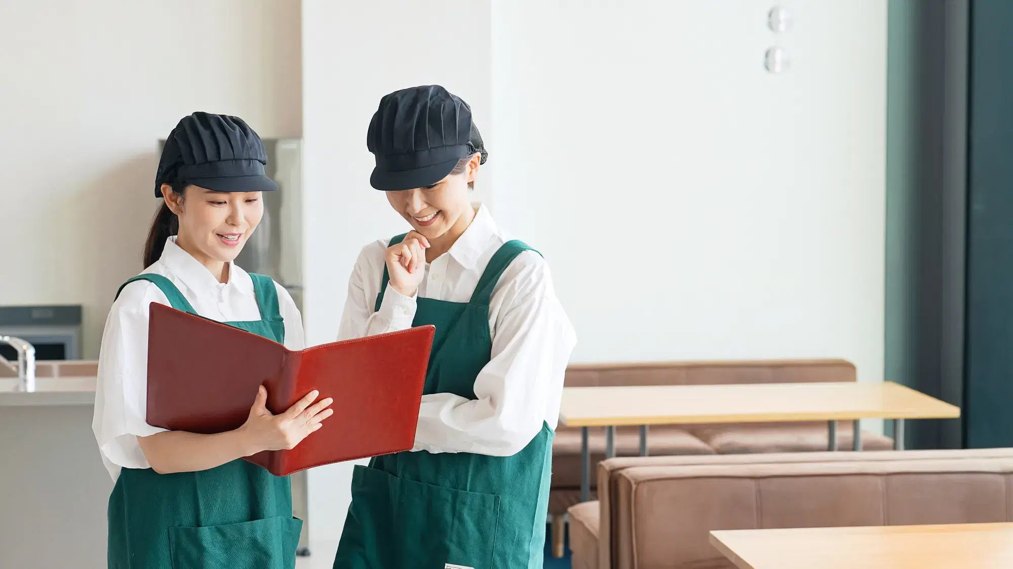 緑のエプロンを着用した片手に赤い本を持つ女性スタッフと赤い本を確認する顎に手を当てている女性スタッフ