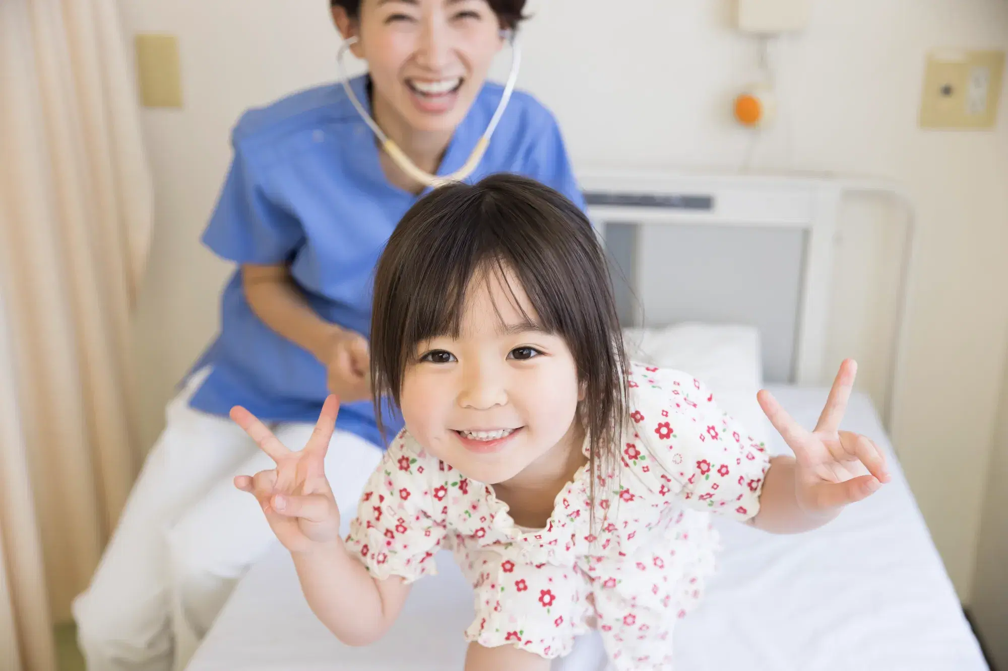 病院のベットの上でピースをしている子どもと青い服を着た笑顔の看護師