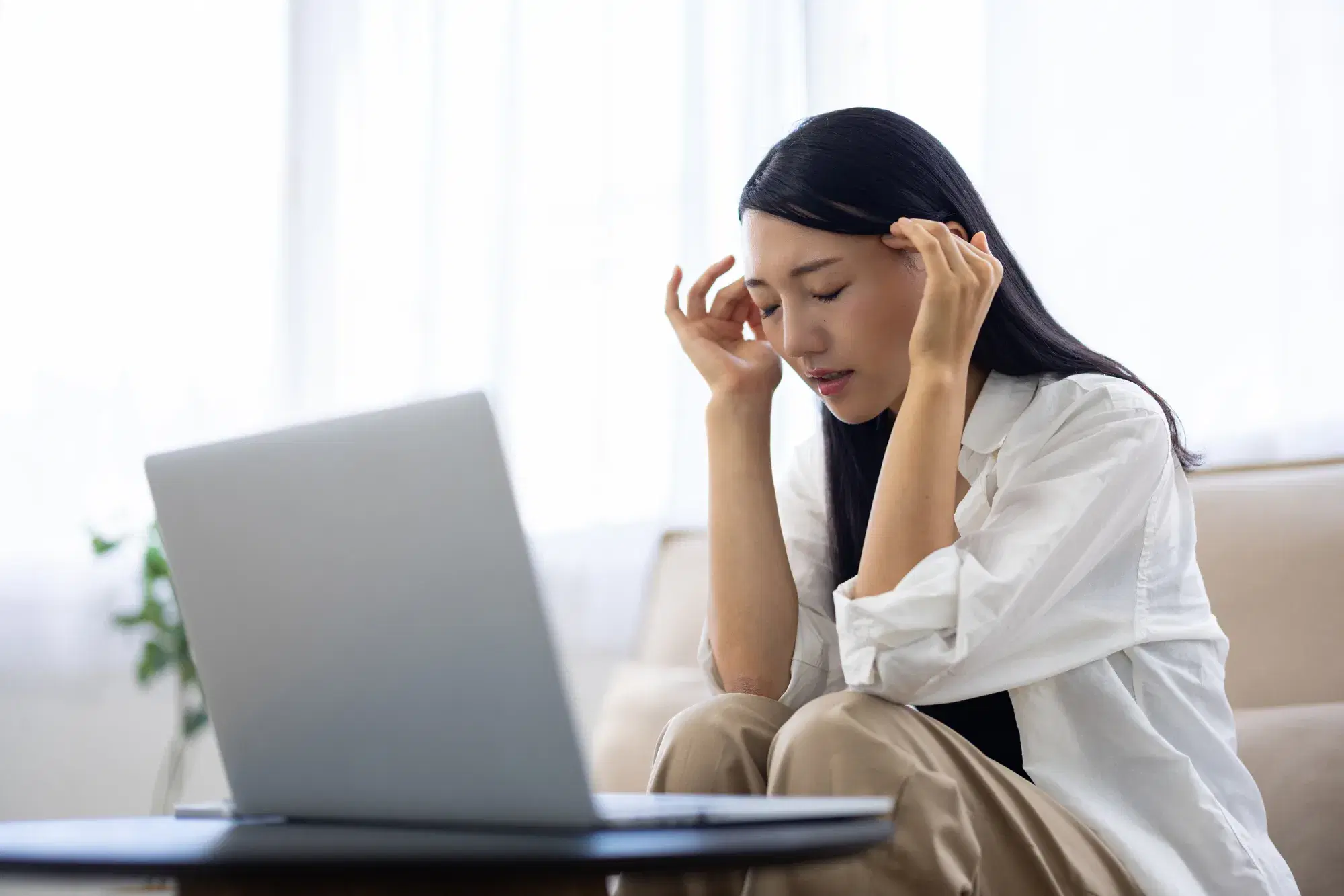 白い服を着た女性がソファーに座りながらこめかみに手を当てている