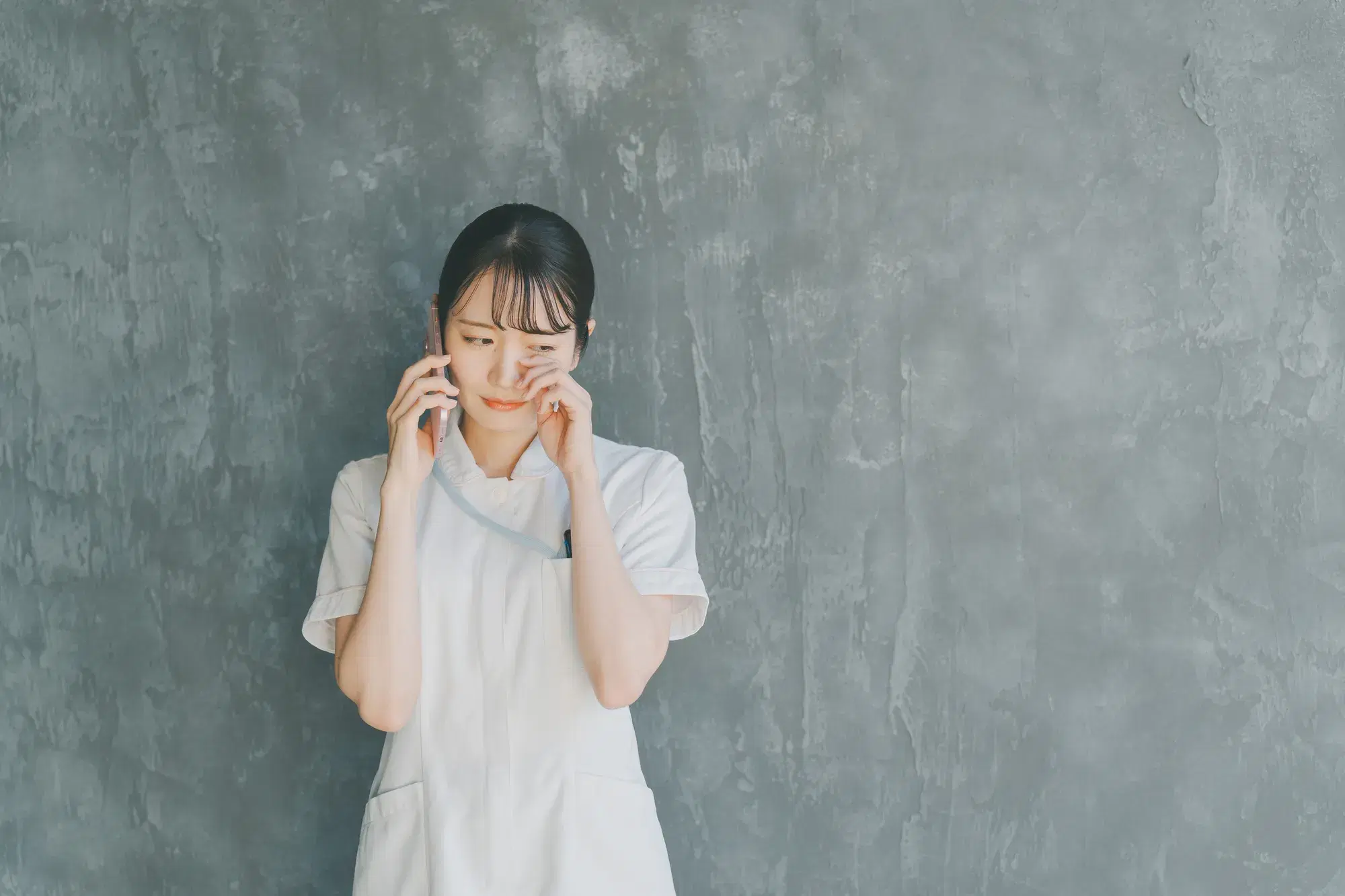 灰色の壁を背にしながら白衣を着た女性が泣きながら電話