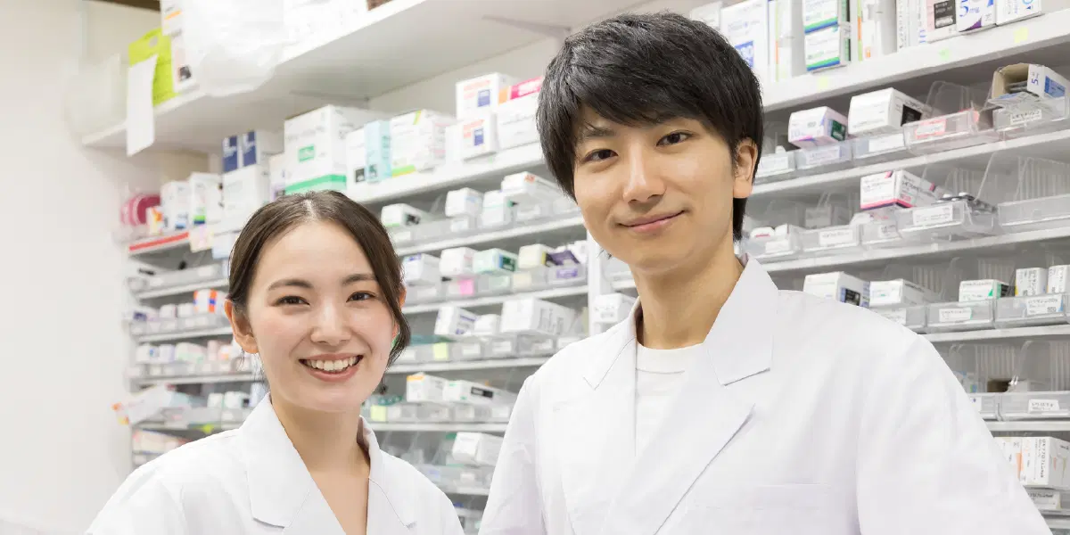 白衣を着た男女が薬剤の入った棚を背景に笑っている