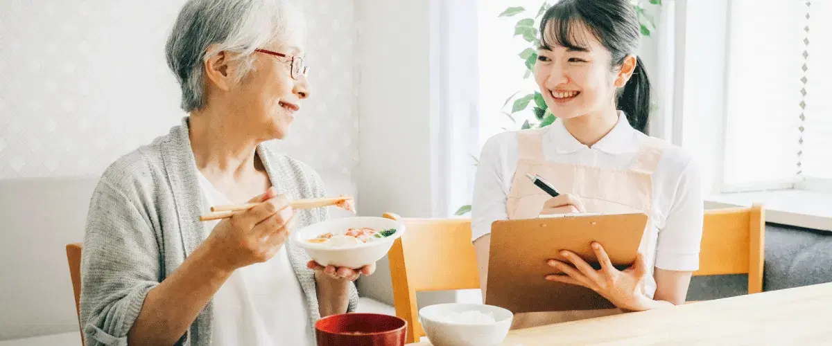 食事中の利用者と笑顔でバインダーにメモを取る職員女性