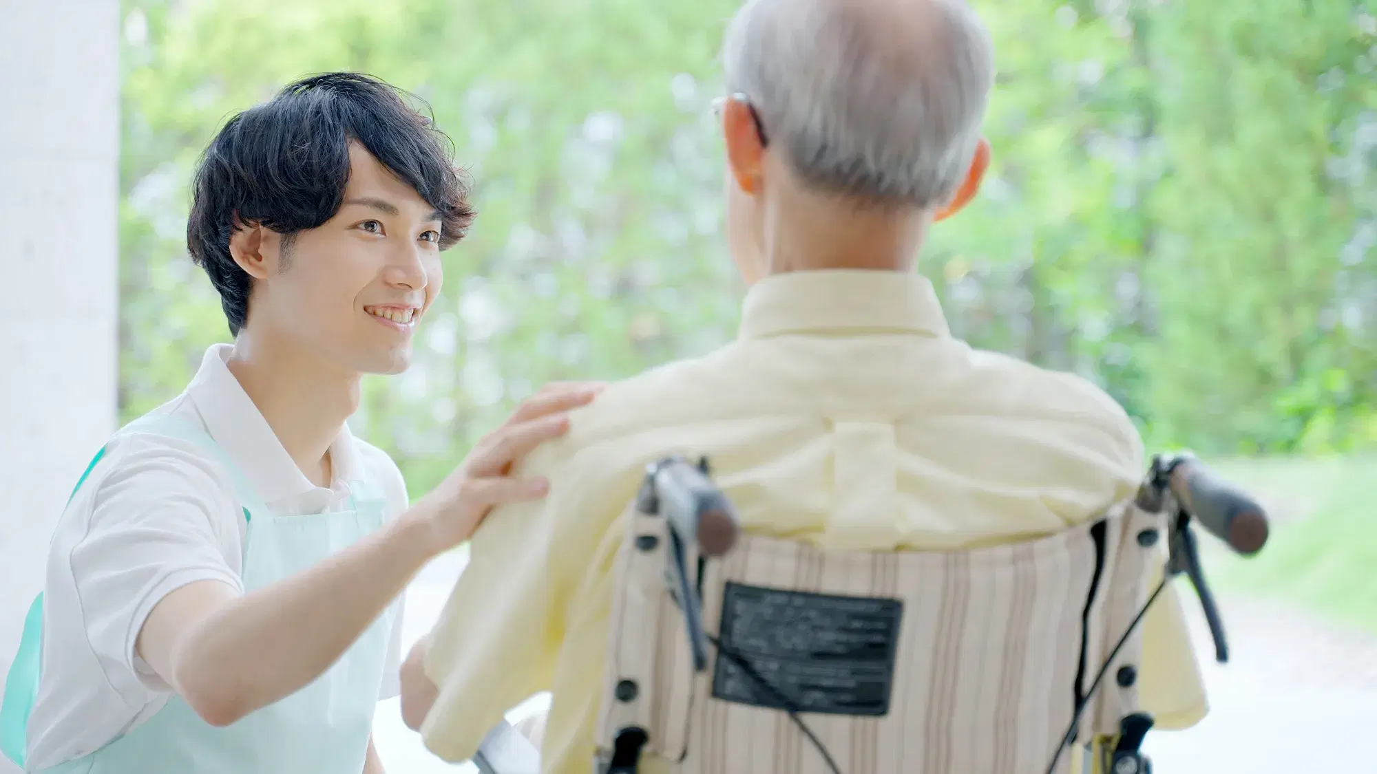 車椅子に乗った黄色の服を着ている男性の左肩を白い服の上に水色のエプロンを着用した男性が掴んでいる