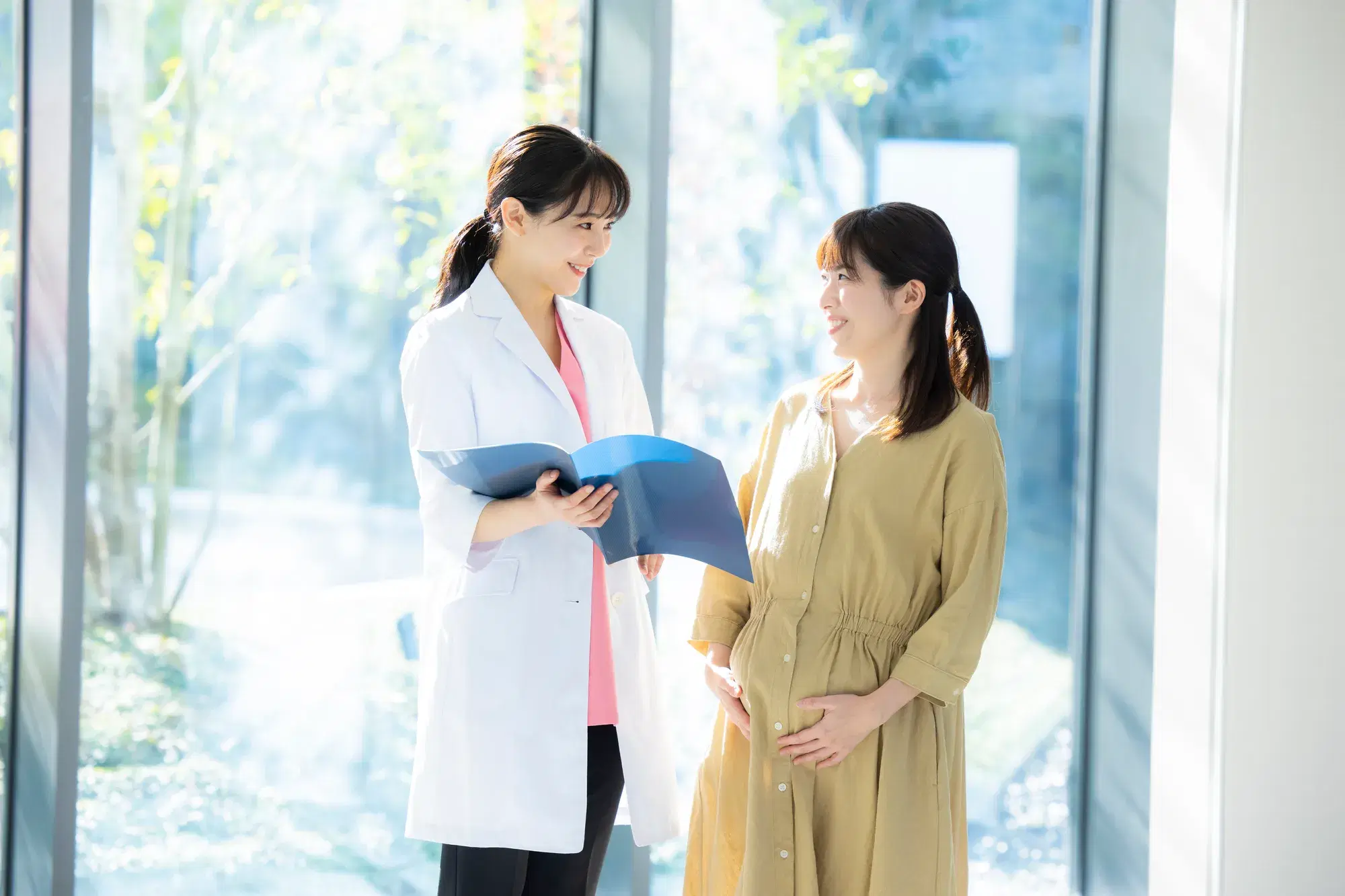白衣の下にピンクの服を着ている女性の医師と、黄土色のワンピースを着た妊婦が窓の近くで話している