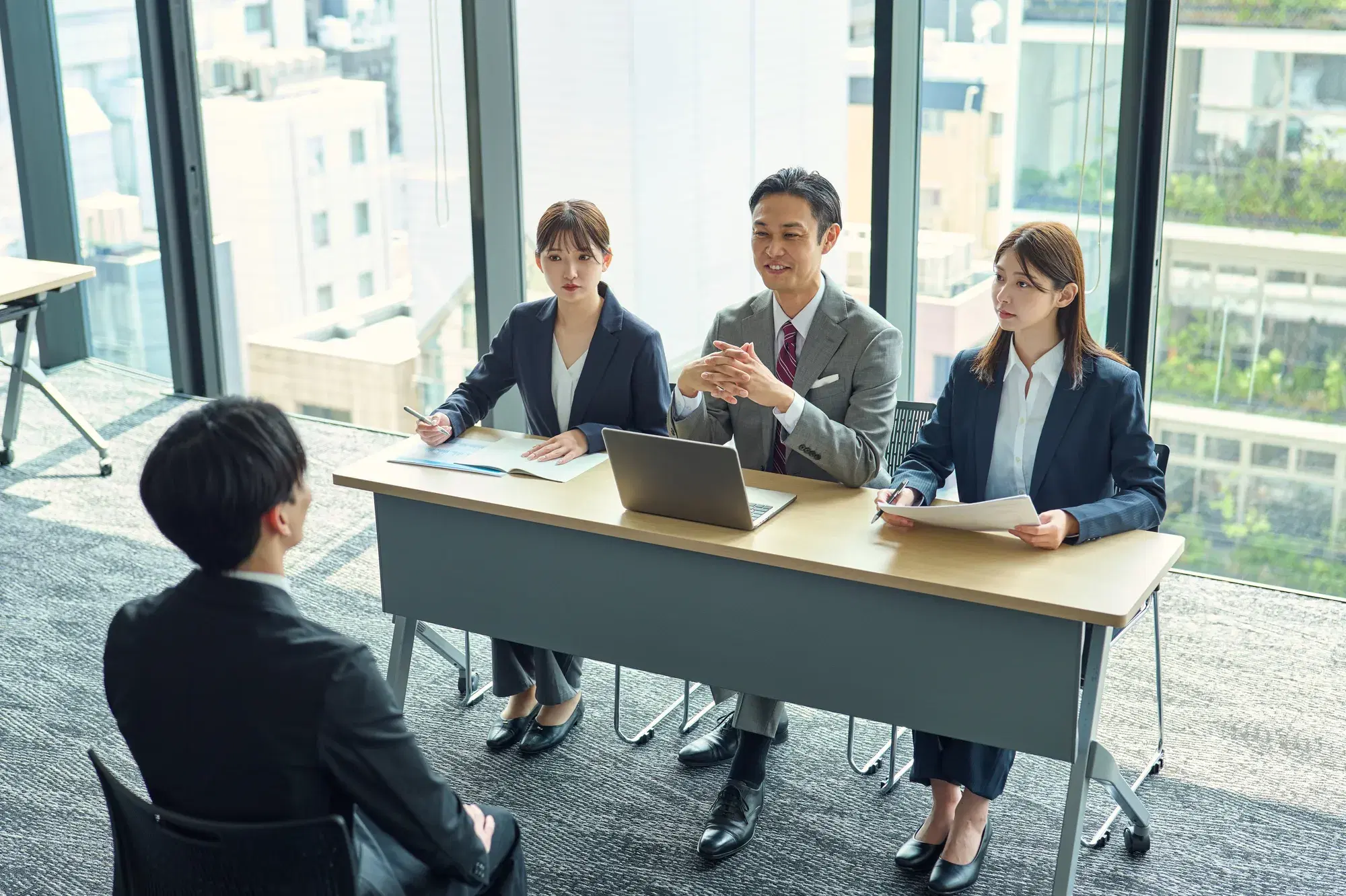 明るいオフィスビルの一室で、デスクを挟んで並ぶ3人の面接官(女性2人、男性1人)と向き合い、面接を受けているスーツ姿の求職者の男性