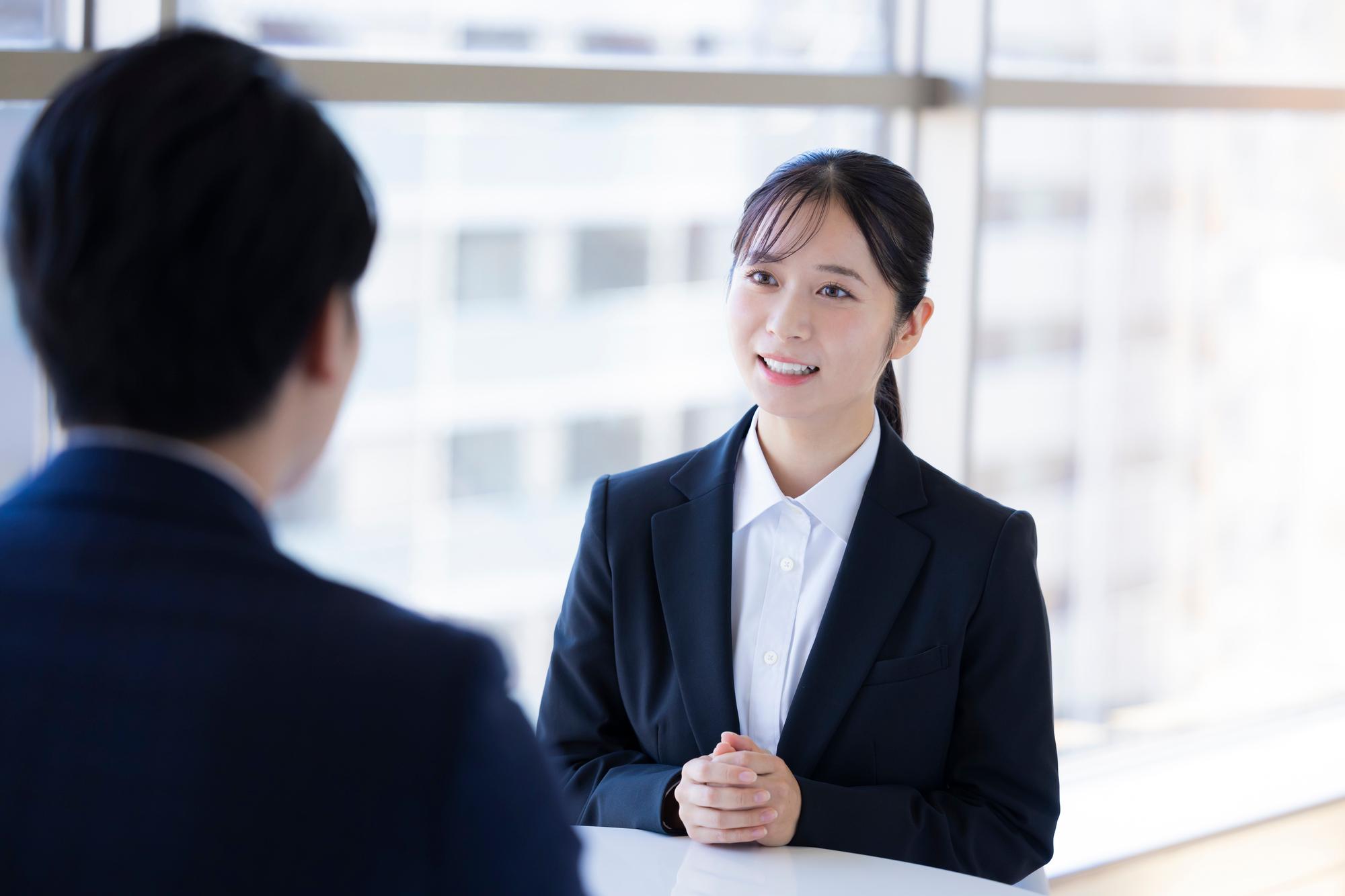 手前に紺色のスーツを着た男性がおり、奥で黒いスーツを着た女性が手を組んで座っている。