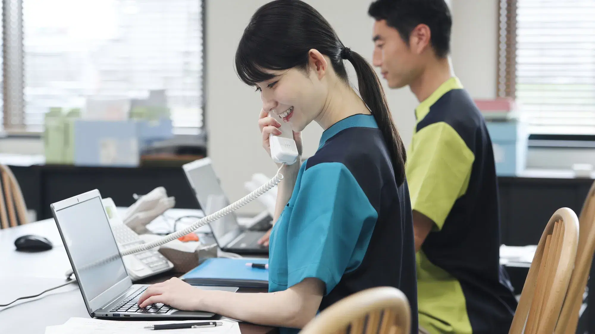 机の上の受話器を取りながら左手でパソコンの操作をする黒色と青色の服を着他女性