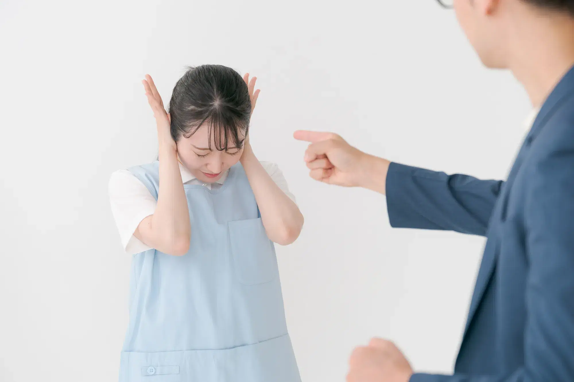 水色のエプロンを着用した女性が男性に指を指して怒られており、女性は耳をふさいでいる