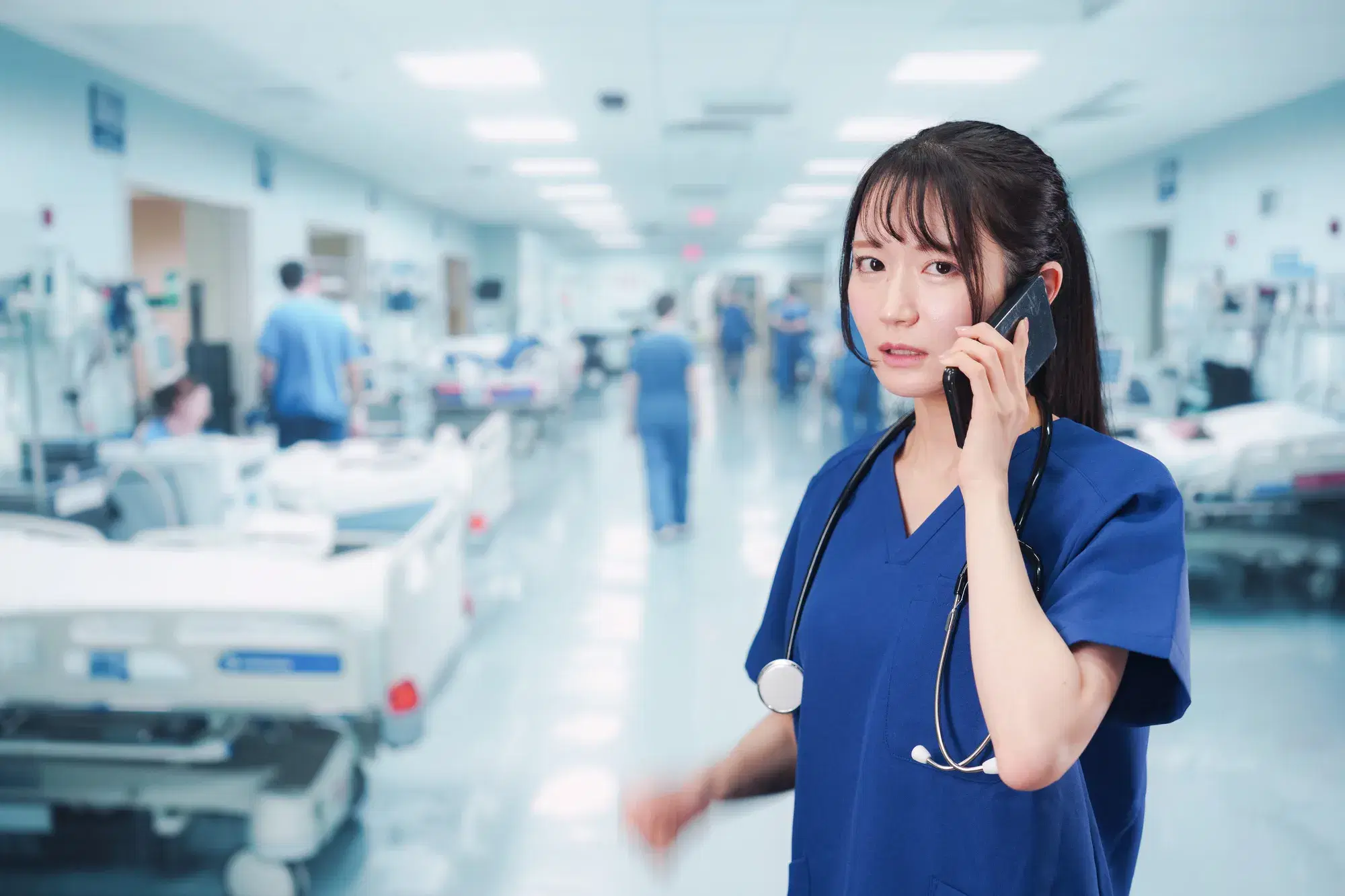 紺色のスクラブを着た女性の医療従事者が、病室内でスマートフォンを耳にあてて話している。