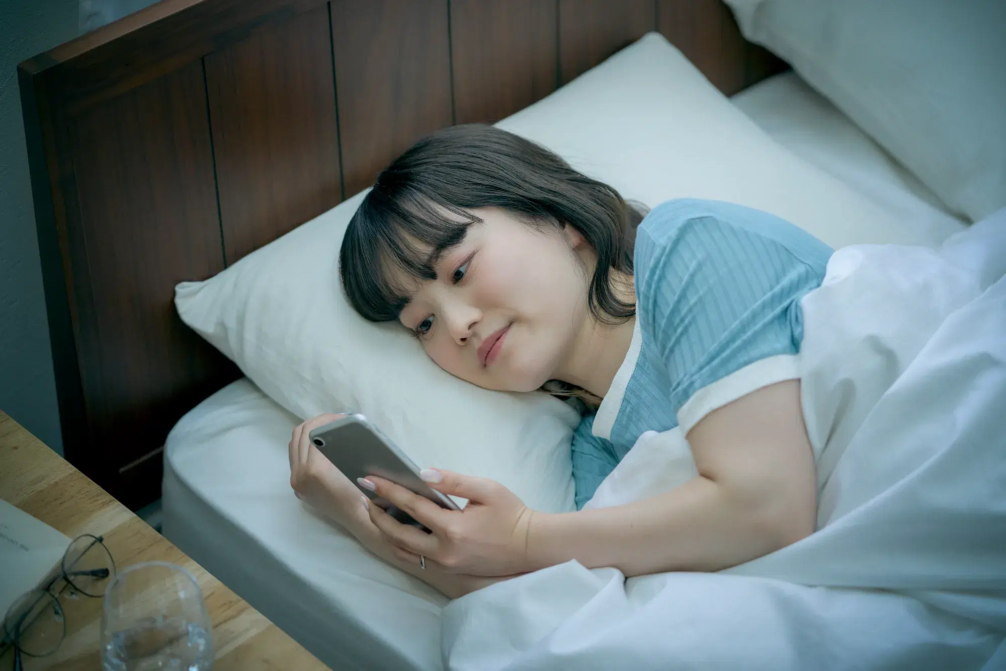 ベットで横になりながらスマホを操作している水色の服を着た女性