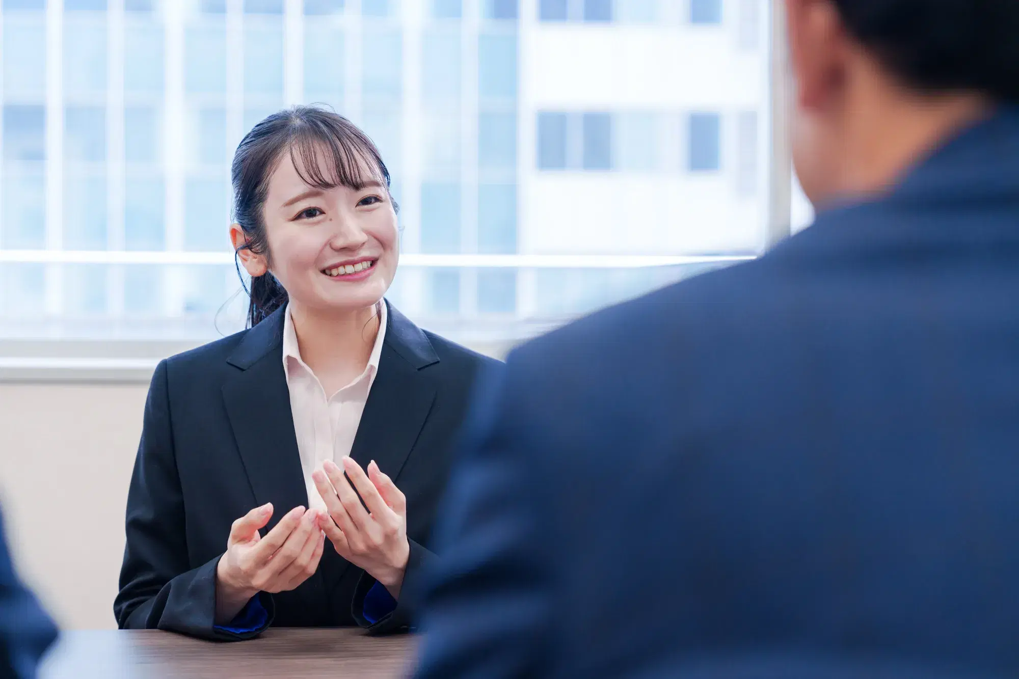 手前に紺色のスーツを着た男性がおり、奥で黒いスーツを着た女性が手を広げて話している。