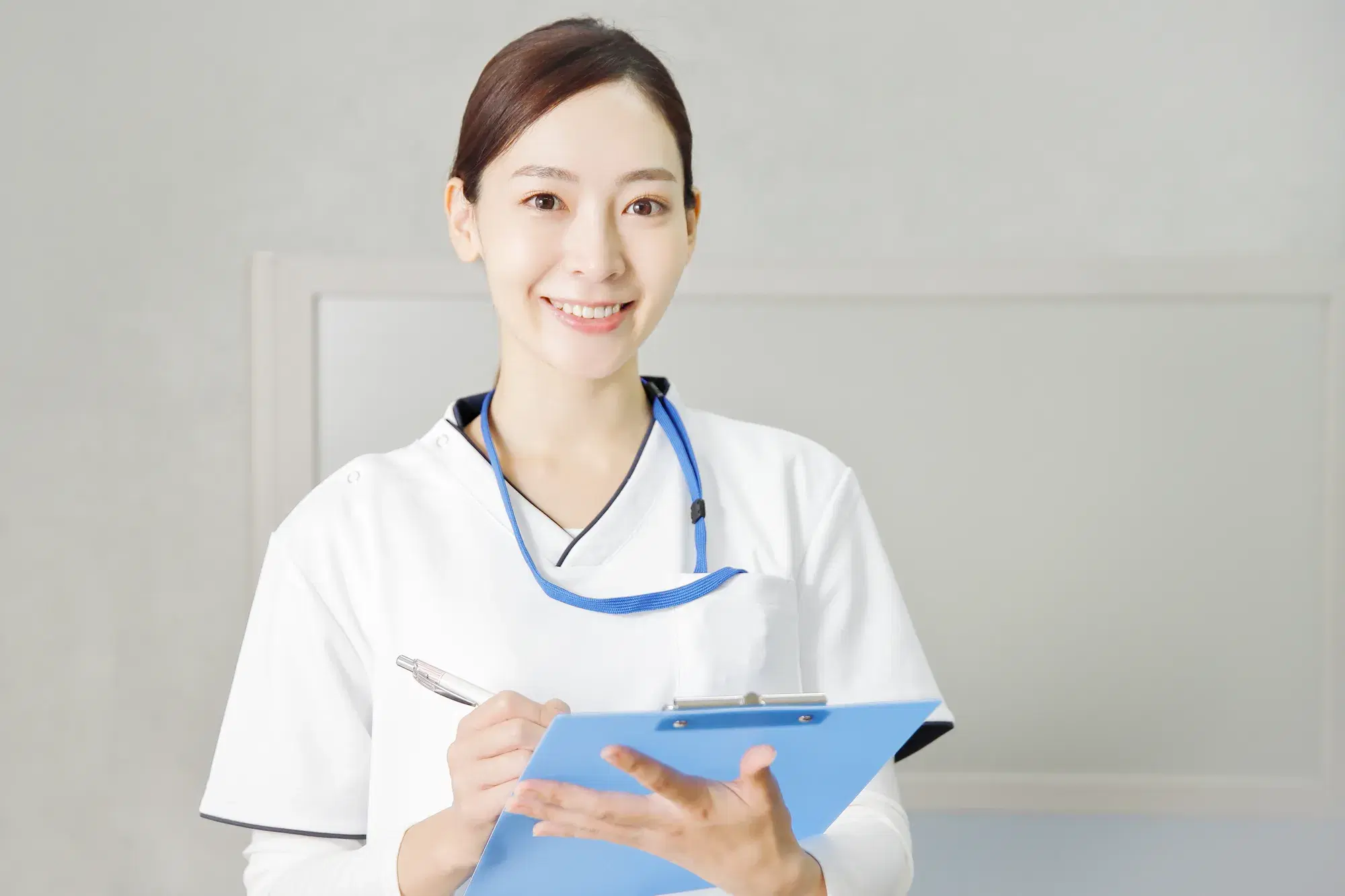白い服を着た女性の医療従事者が、青いバインダーとペンを持って正面を向いている。