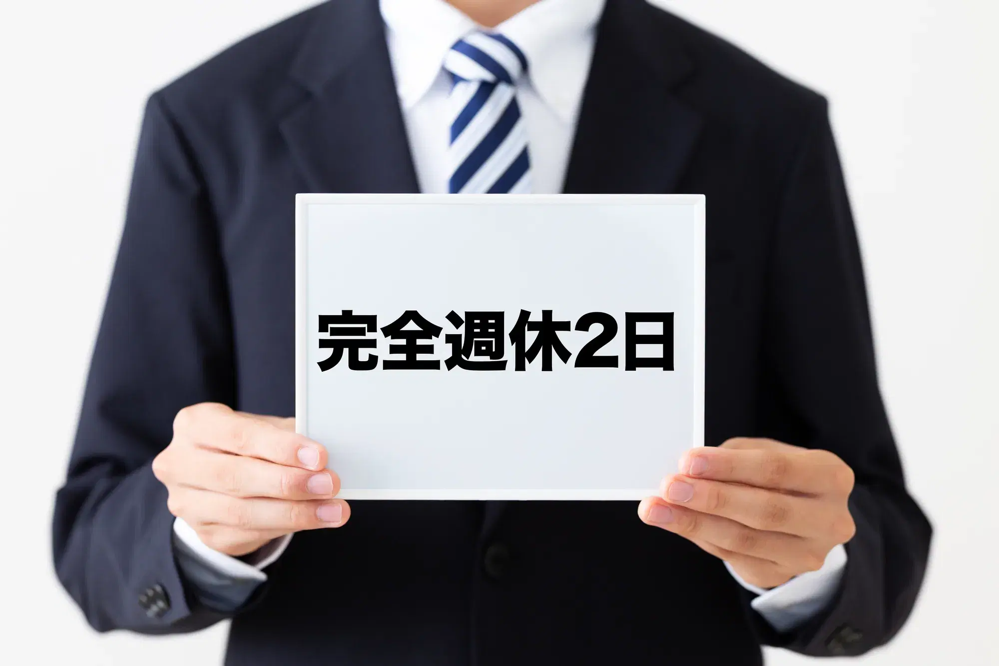 黒い服を着た男性が「完全週休2日」と書かれたボードを両手で持っている。