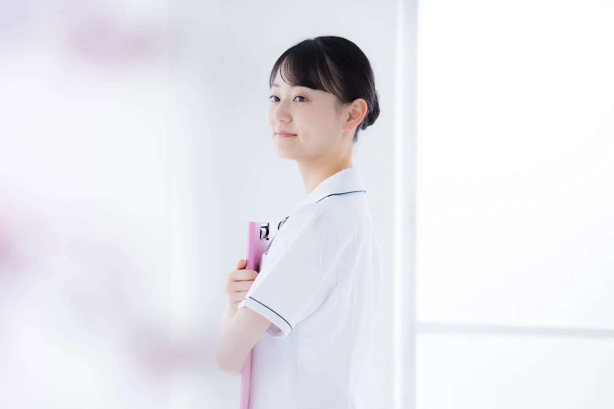白い服を着た女性の医療従事者が、ピンクのバインダーを胸に抱えて立っている。