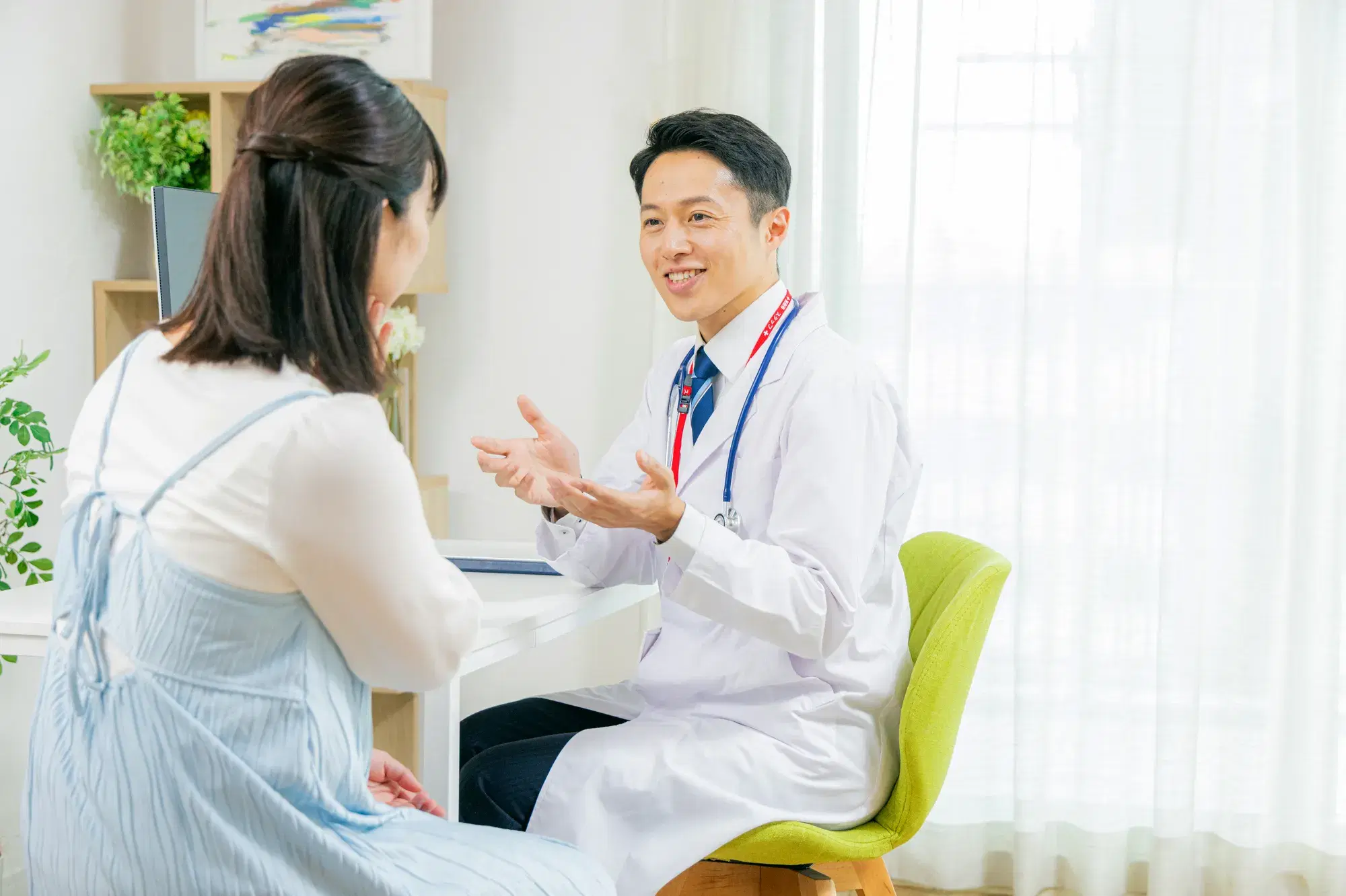首から赤色のストラップと黒い聴診器を下げている白衣を着た男性が水色のワンピースを着た女性に説明