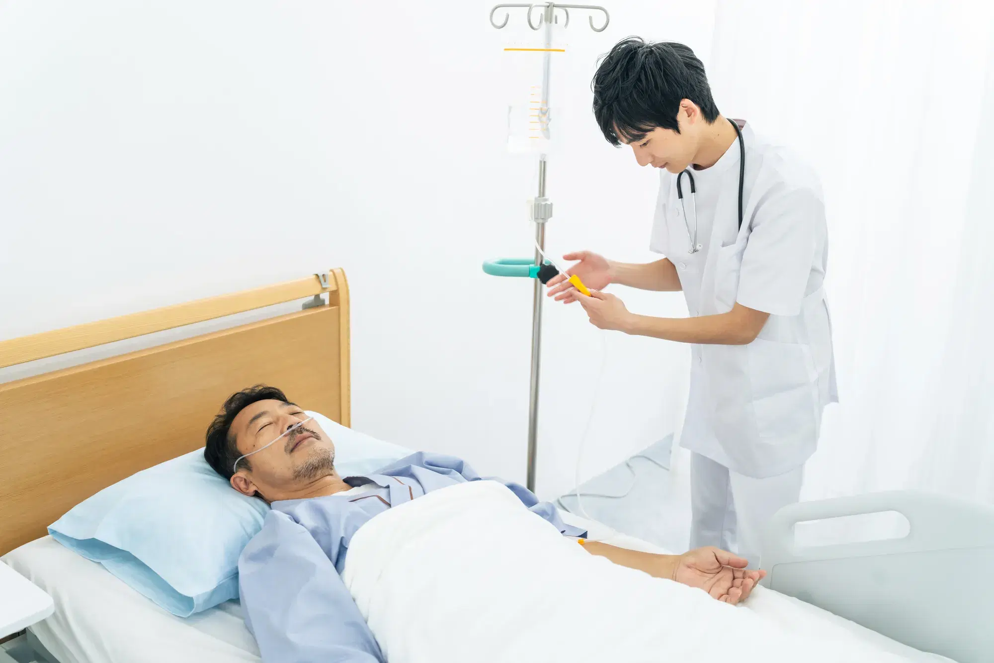 青い服を着た患者がベッドに横たわって点滴を受けており、白衣を着た男性が点滴を触っている