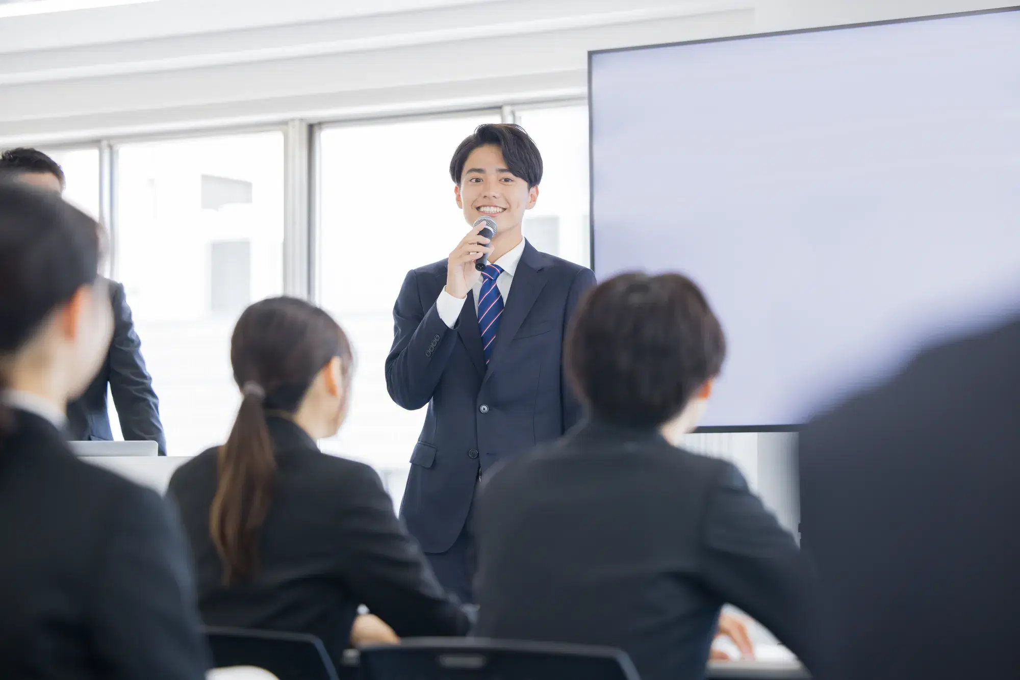 複数の社員の前でスーツを着た男性がマイクを持って発表