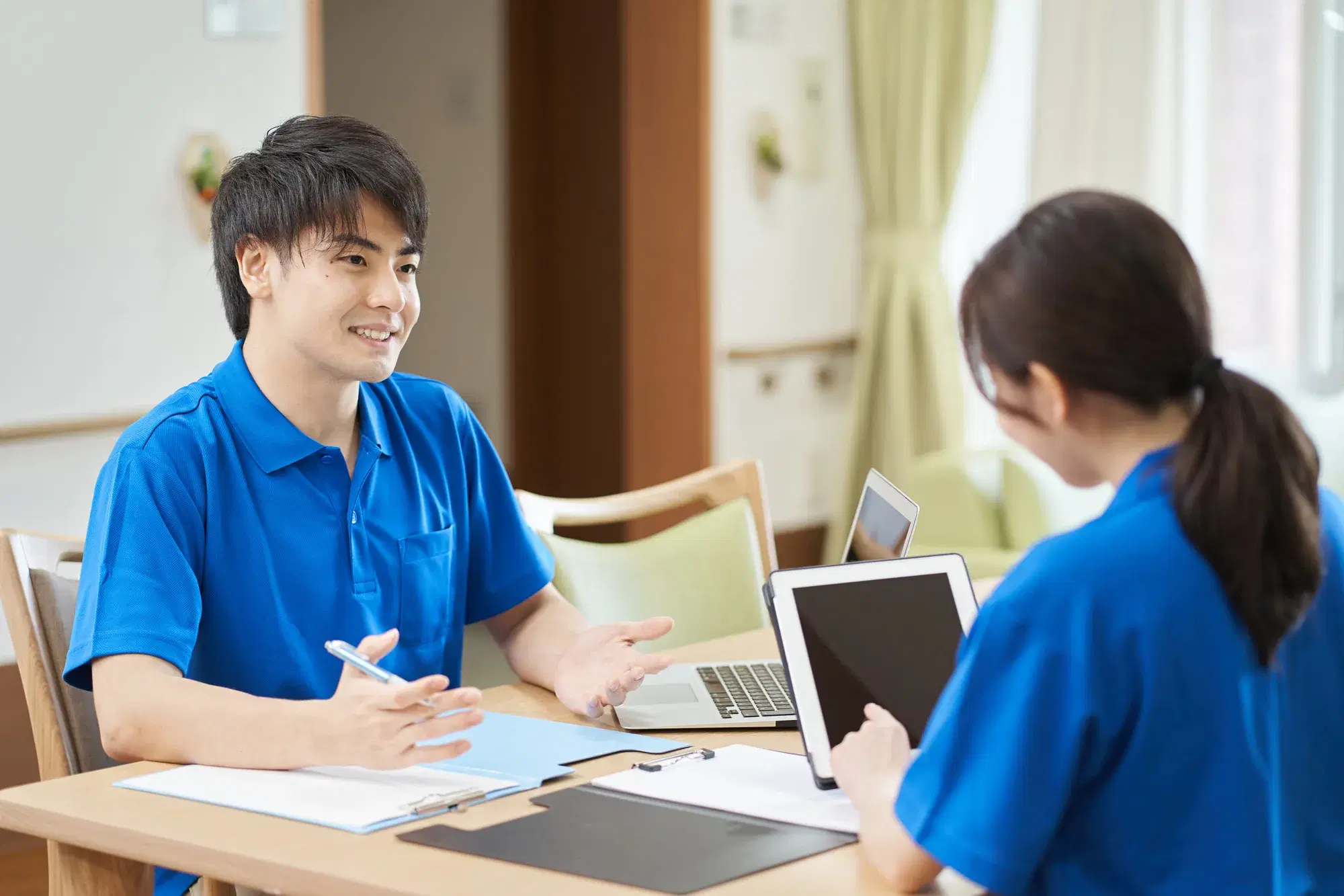青い服を着た男性と女性が机に書類やタブレットを置いて話し合っている
