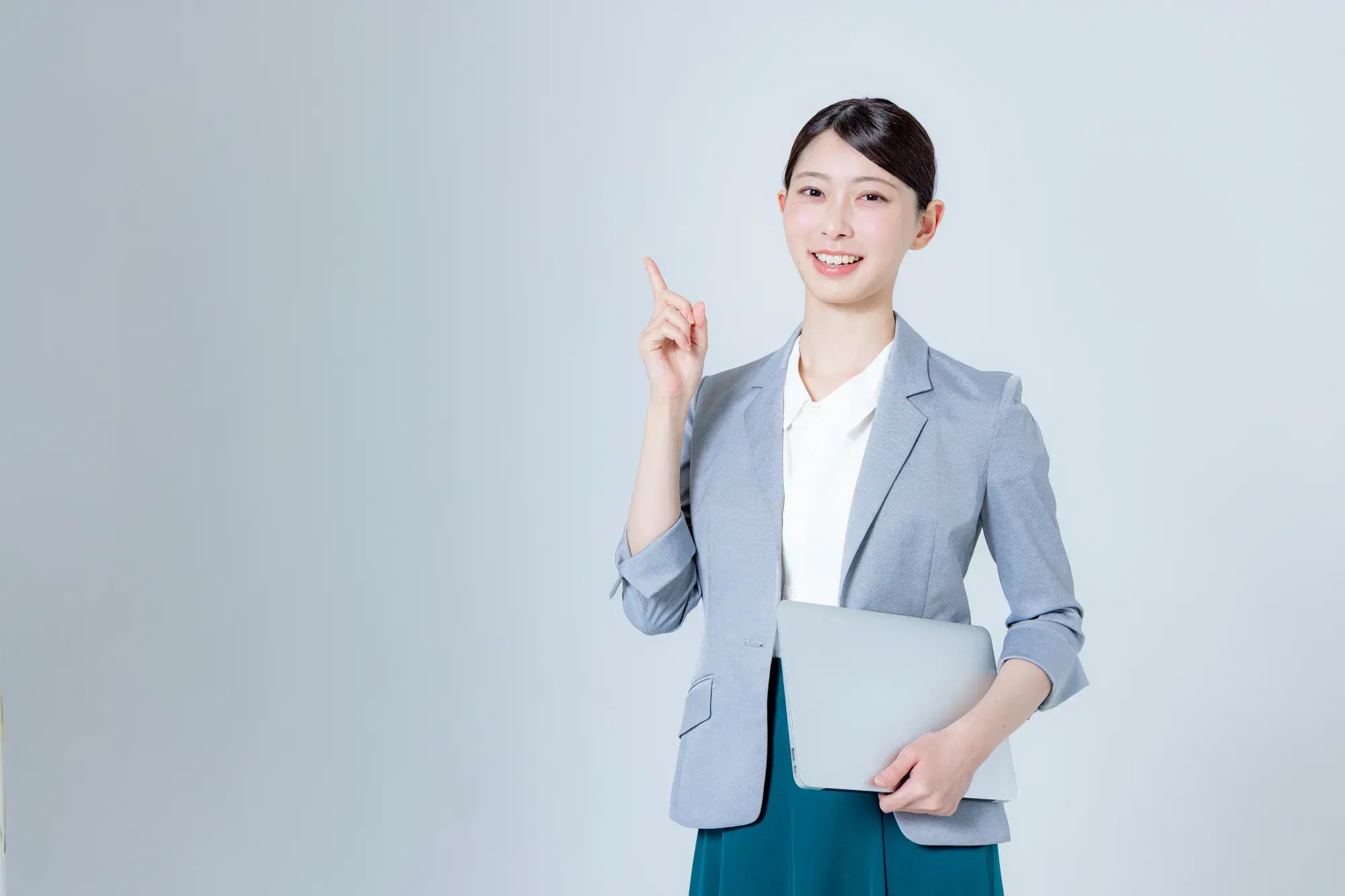 グレーのジャケットを着た女性が顔の横で右手の人差し指を立て左手にはパソコンを持っている