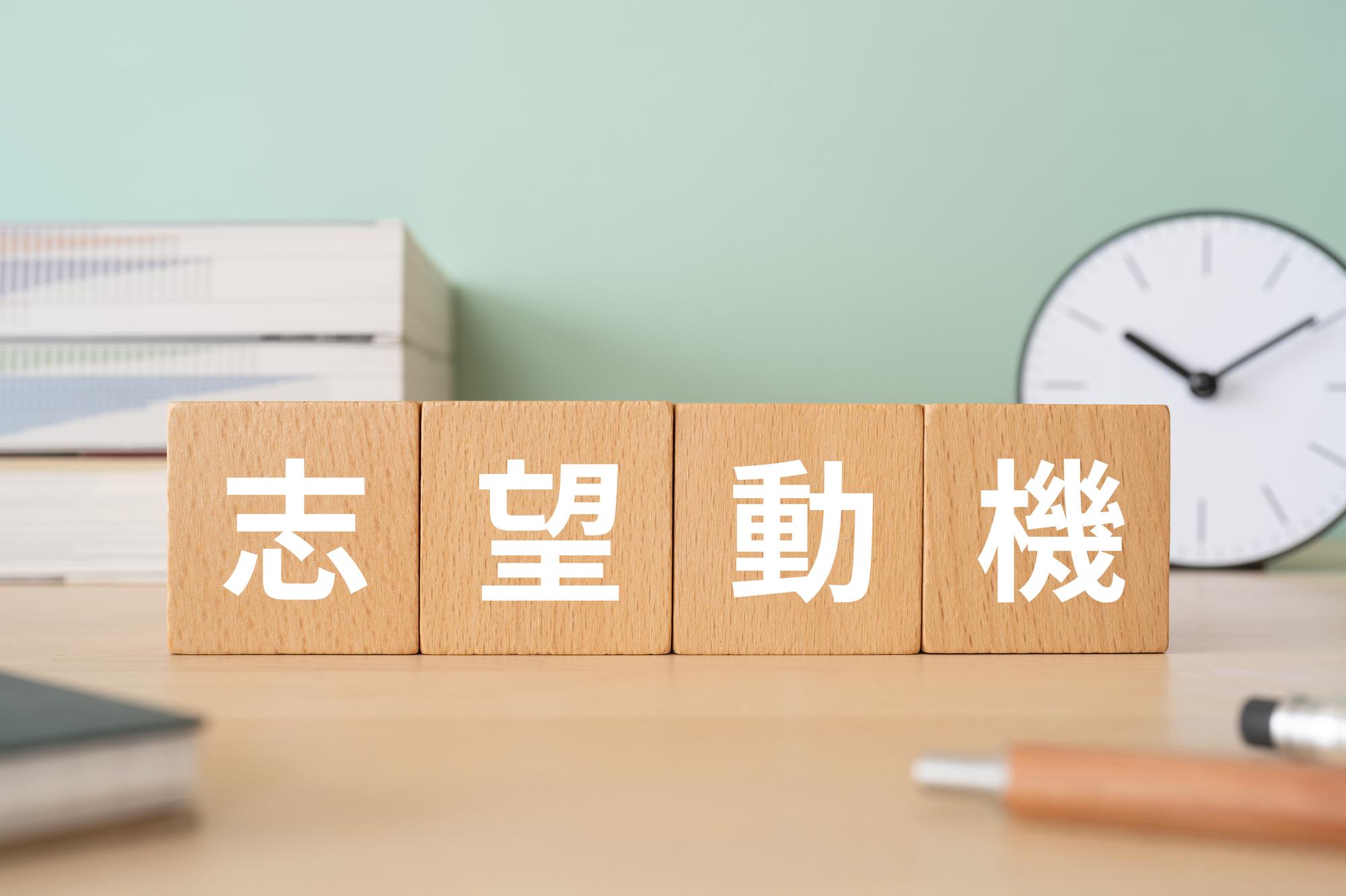 「志望動機」と書かれた木製ブロック、積み重なった本、置き時計、ペンが置かれている様子が写っている。