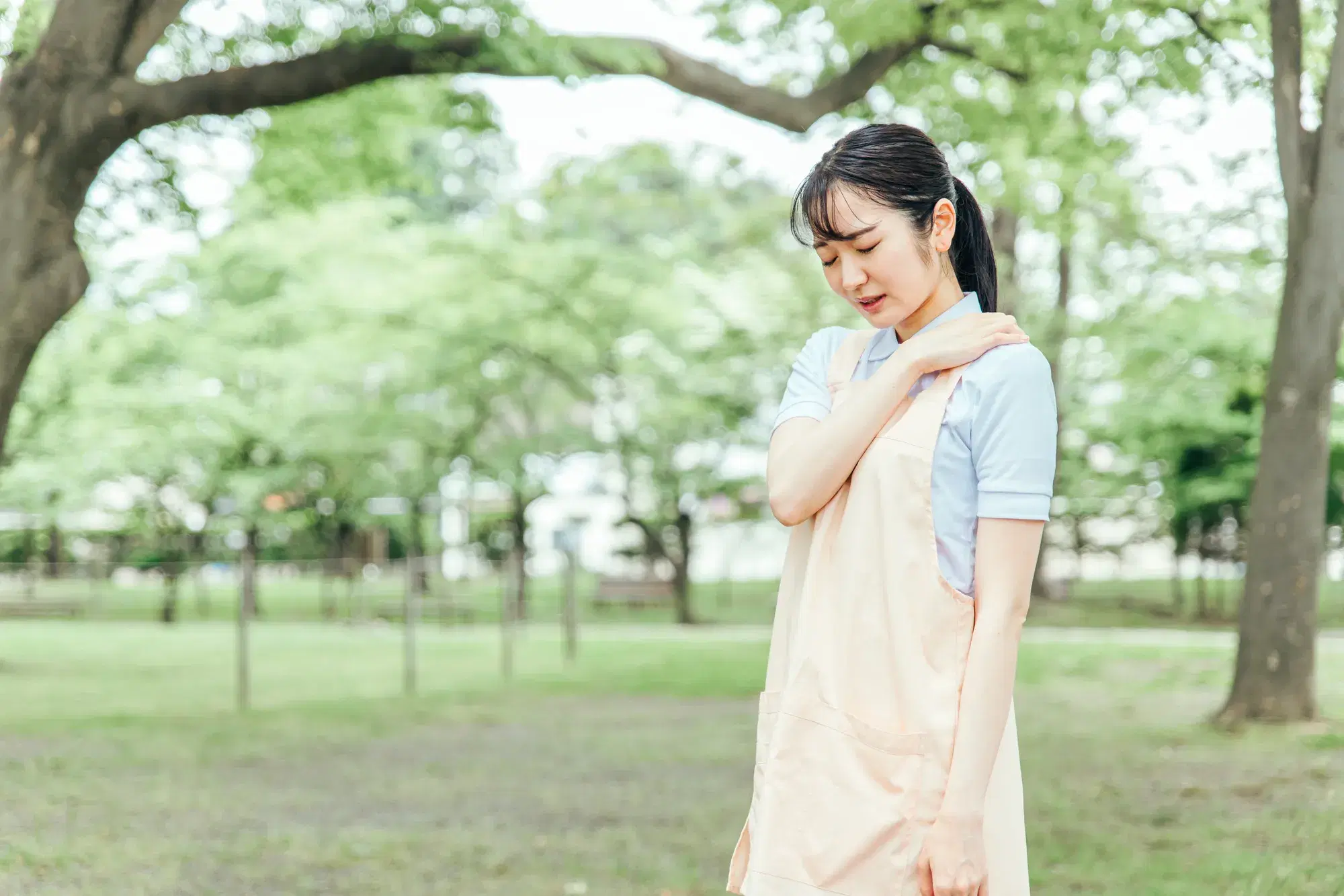 公園でベージュ色のエプロンを着用した女性が左肩に右手を乗せて俯く
