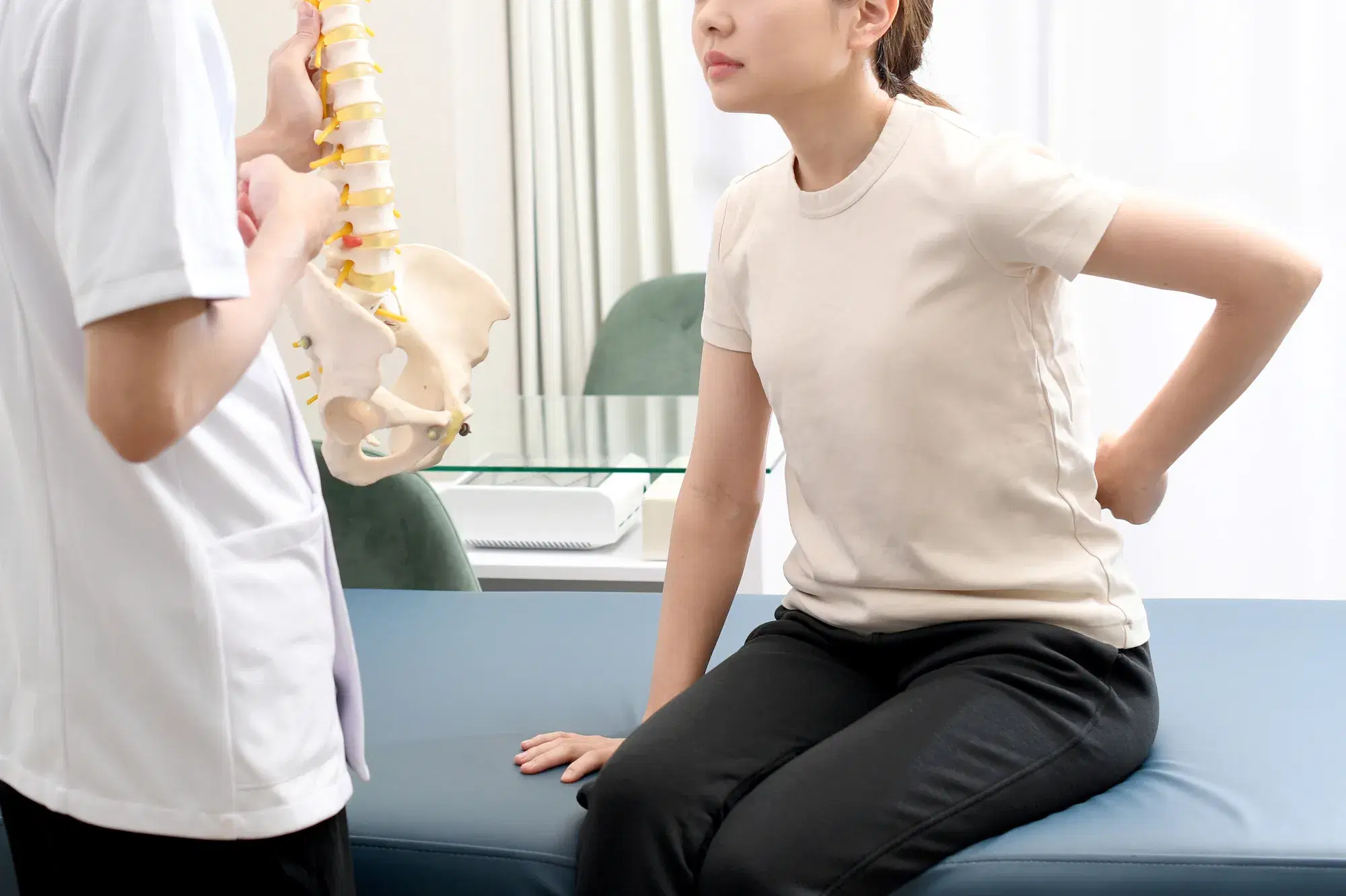白い服に黒いズボンを履いている女性が腰の痛みを訴えているのに対して白い服を着た整体師が模型を使って説明している