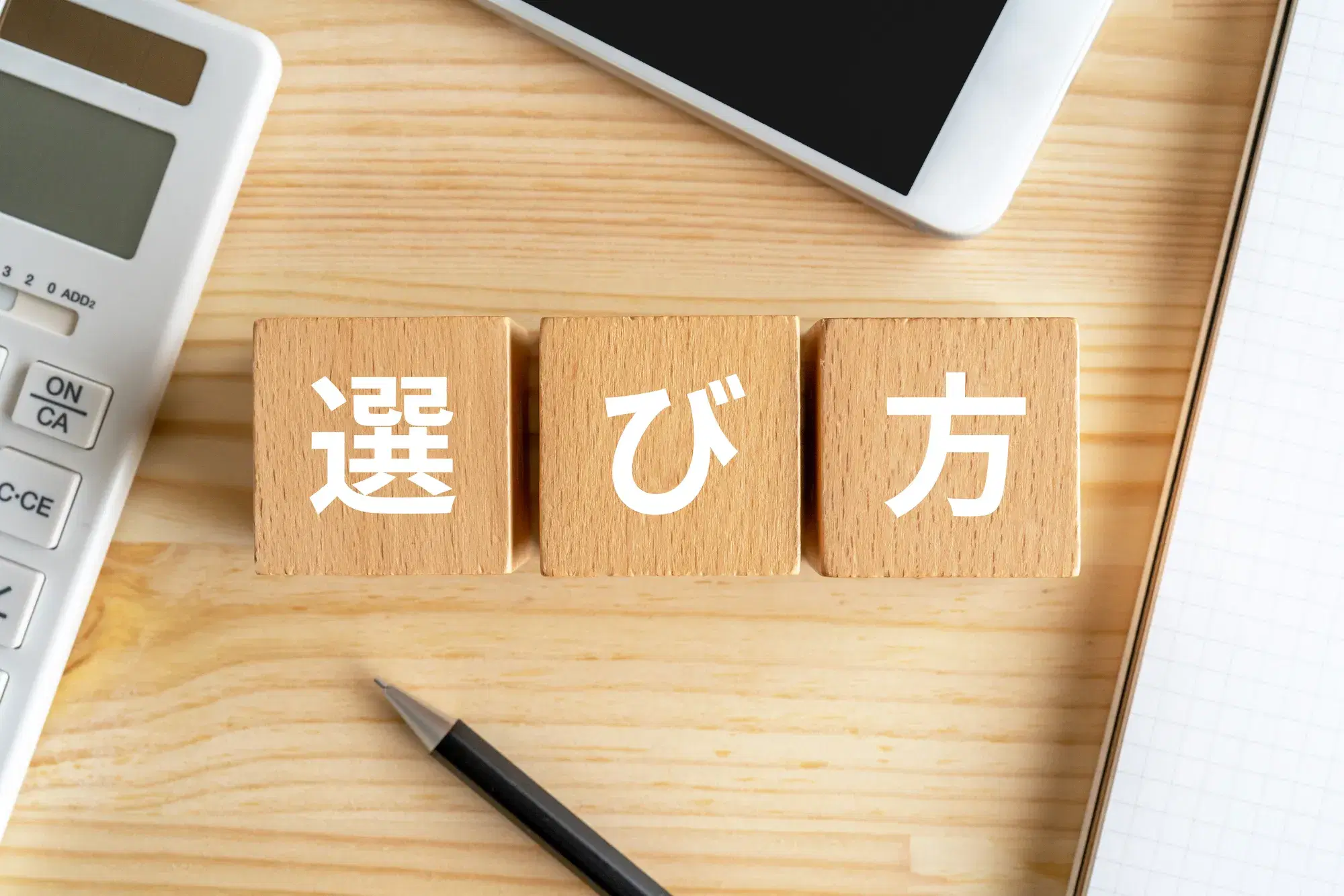 白い文字で選び方と書かれた木製の積み木の周りが鉛筆や電卓やスマホ、紙で囲まれている。