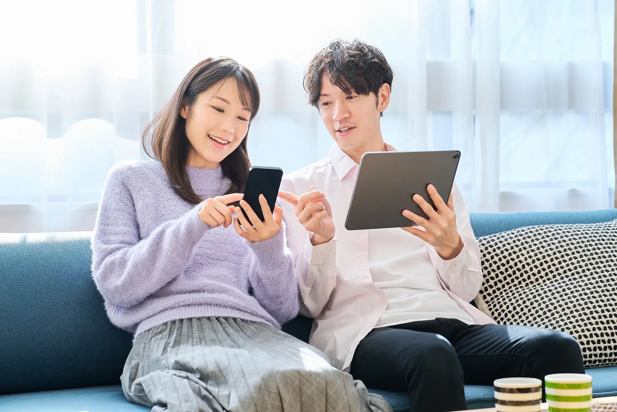 紫の服を着ている女性がスマホを見ており、その隣にいる白いシャツを着たタブレットを持つ男性が女性のスマホを指さしている。